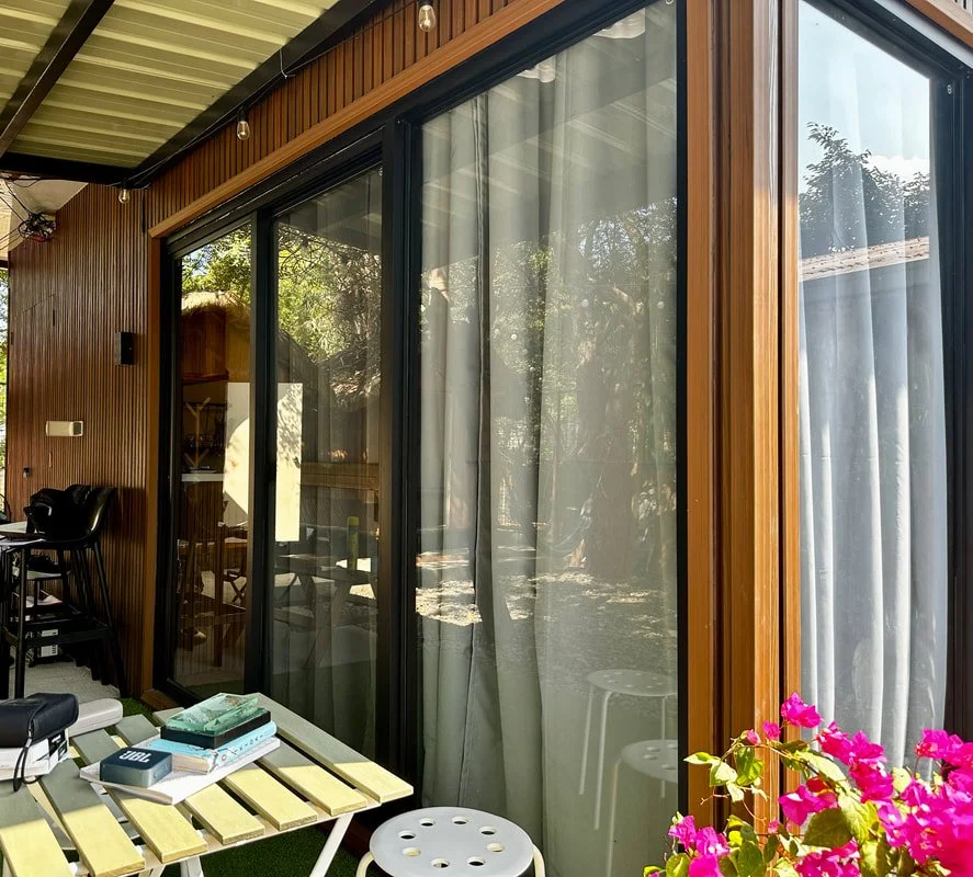 A modern house with wooden siding and large sliding glass doors, outdoor patio with books and colorful flowers.