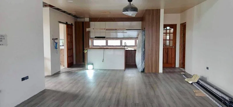 Empty kitchen and living area with gray wood flooring, white walls, and wood accents, under construction or renovation.