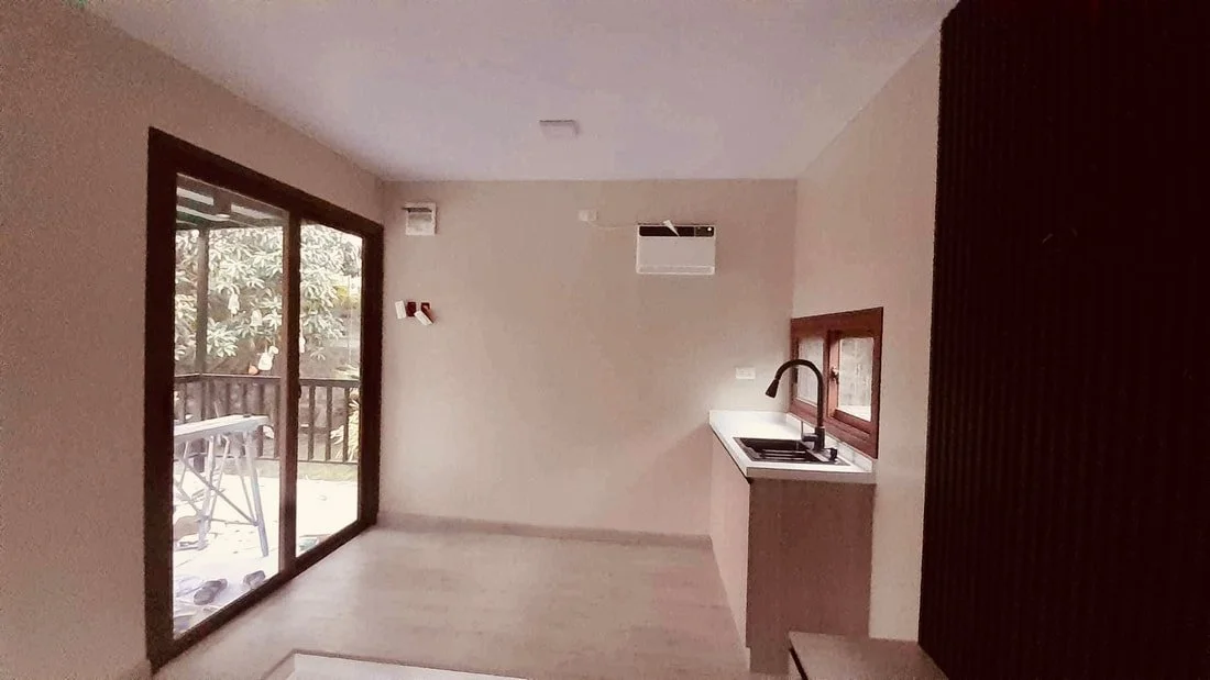 Empty room with a sliding glass door leading to a balcony, small kitchen area with a sink and window, wall-mounted air conditioner, and a wooden sliding door to the right.