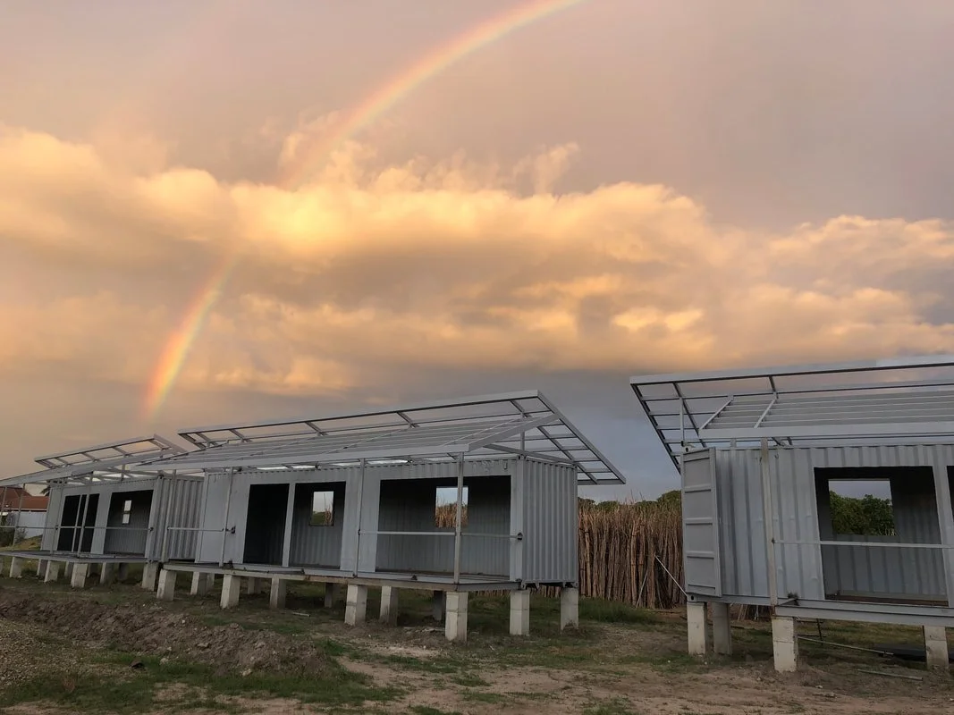 There are two house frames built from shipping containers with metal roofs, set on elevated concrete piles. The sky above has clouds with a faint rainbow.