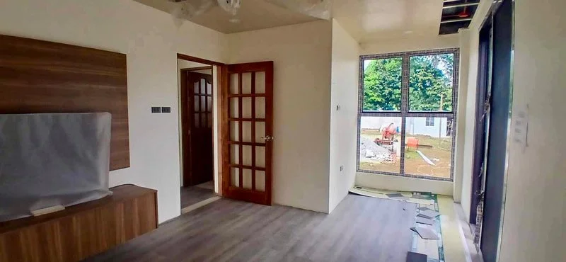 Empty room with wood flooring, a wooden bench, a door with glass panes, and a large window showing an outdoor area with trees and construction materials.
