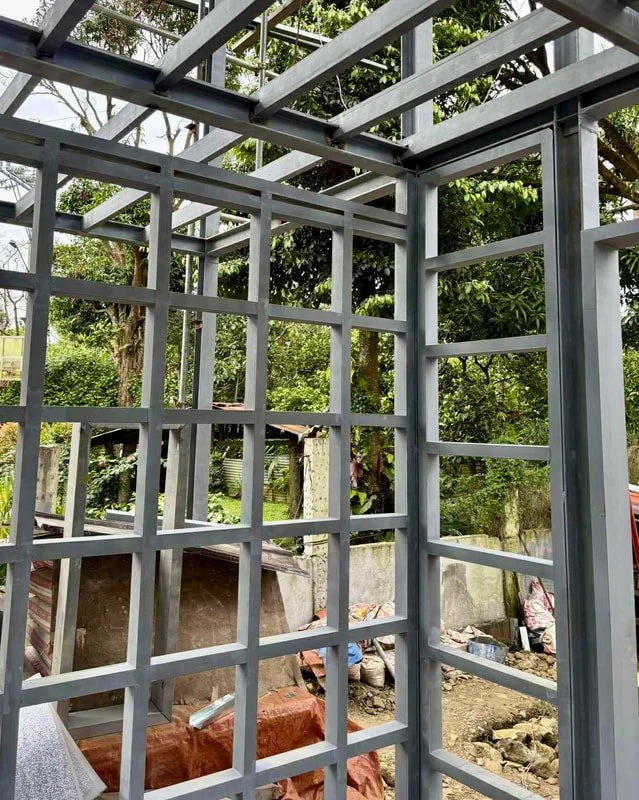 Metal framework for a building under construction, with a grid pattern on the walls and ceiling, outdoors with trees and construction materials visible in the background.