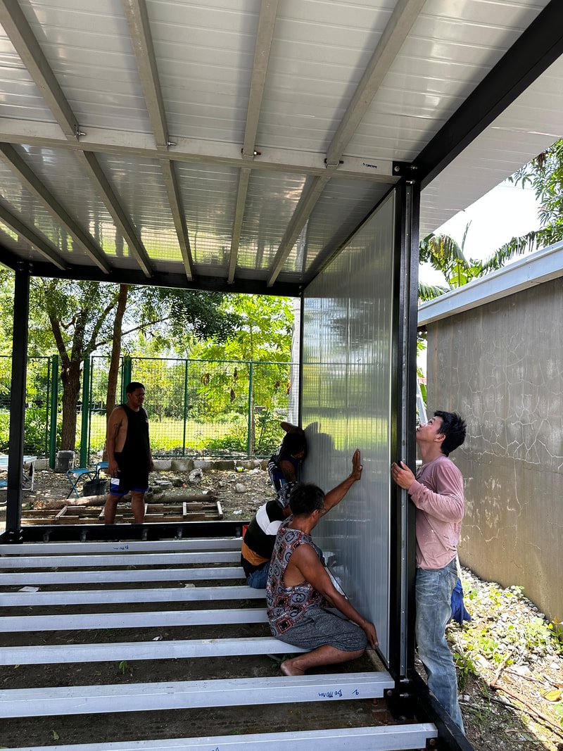 Four people are assembling a metal structure outdoors. One man is holding a vertical panel in place, while a woman on the ground supports it. Two other people are in the background, working near the ground.
