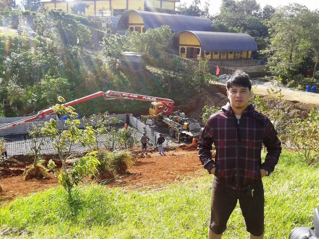 Young man standing on grassy slope with construction site and colorful houses in the background.