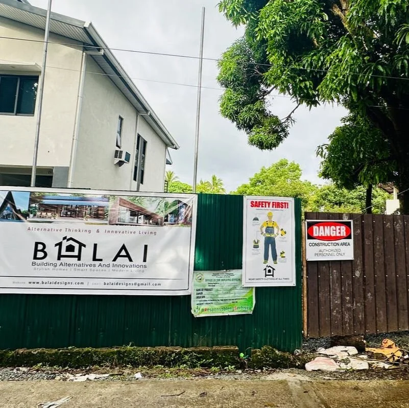 Signs and banners on a construction site fence, including a safety notice, a danger warning, and an advertisement for a building company called Balai Designs, with a partially visible building and trees in the background.