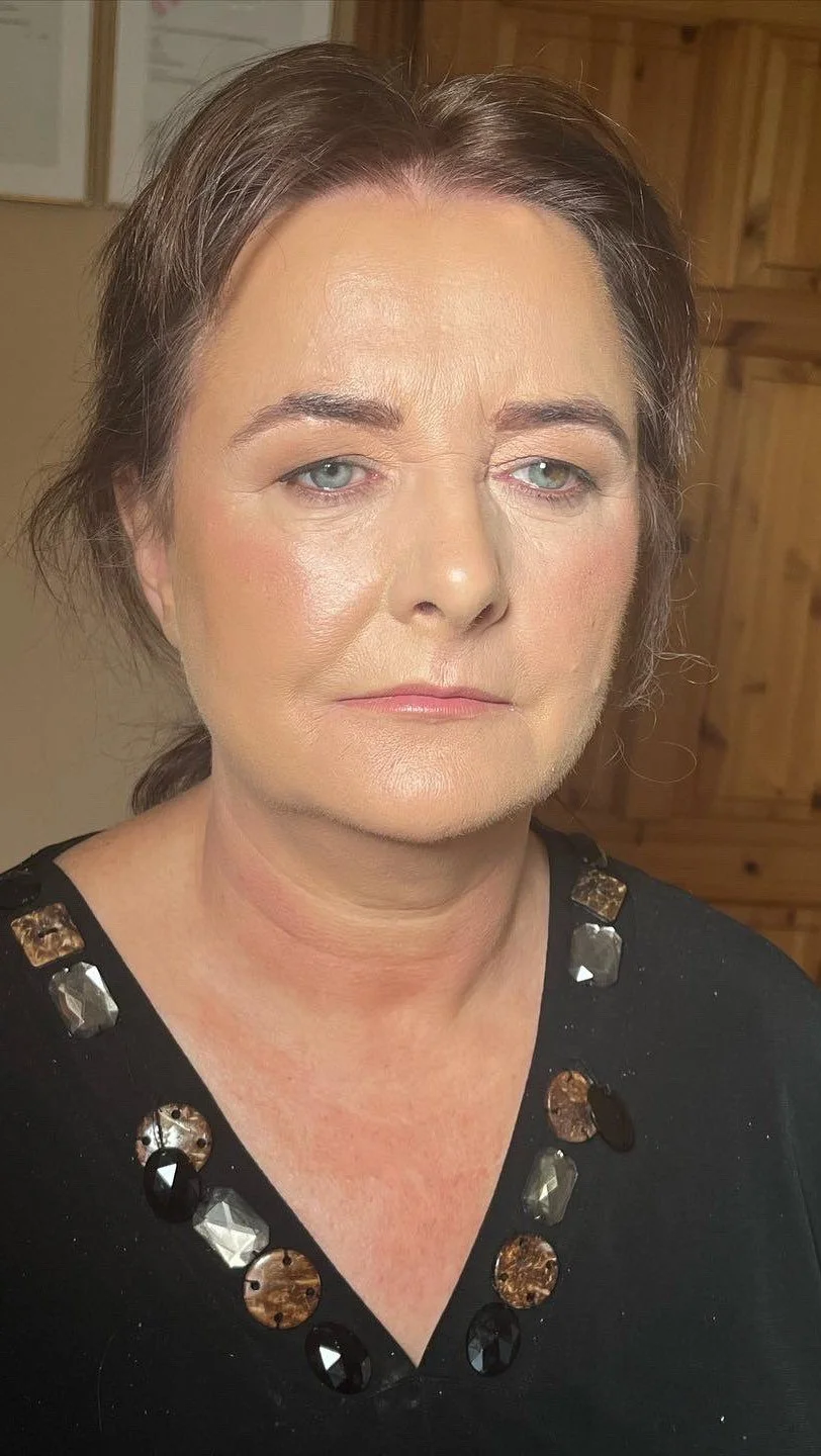 A middle-aged woman with light skin, short brown hair, and blue eyes. She is wearing a black top with decorative beads around the neckline, standing in a room with wood-paneled walls.