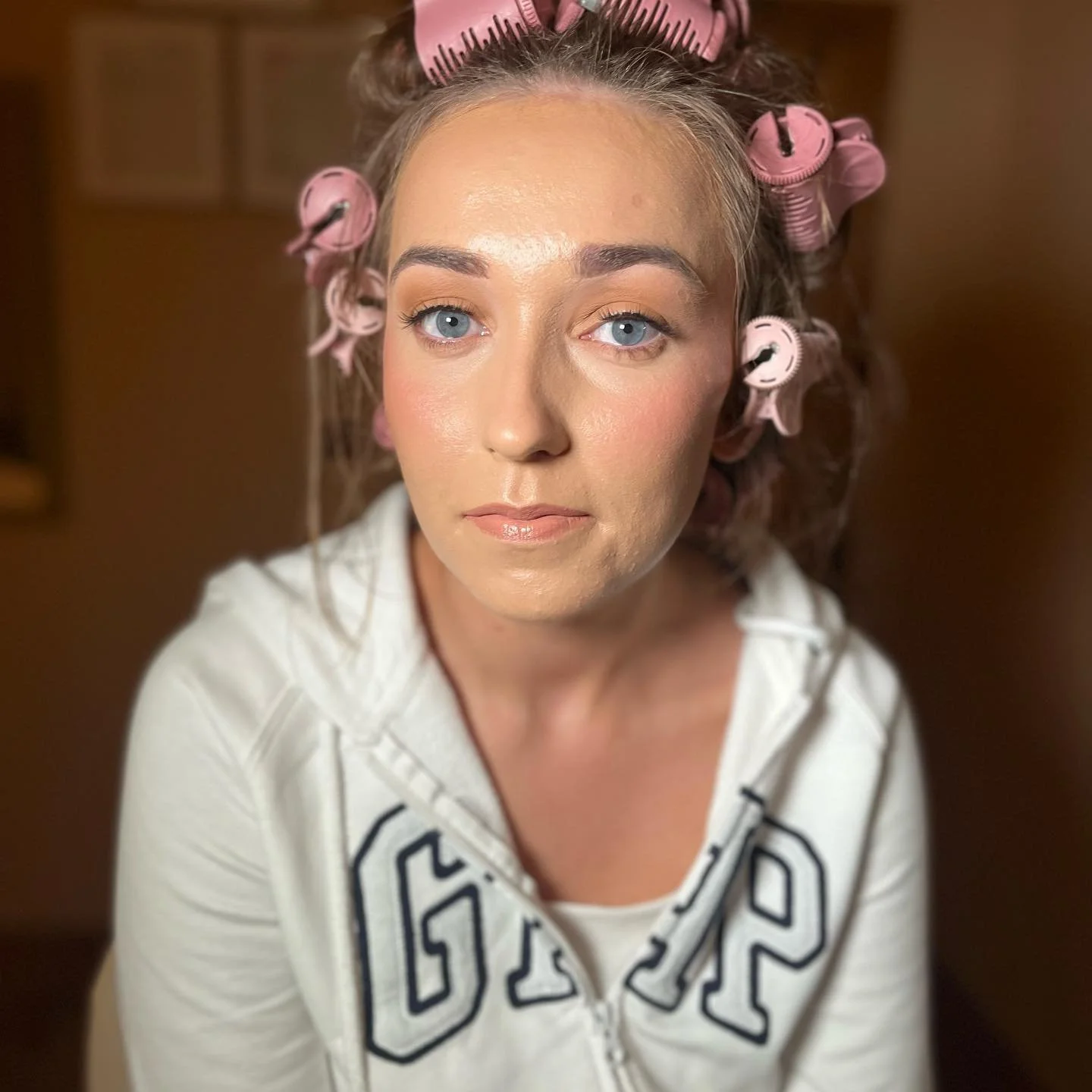 Woman with grey hair in pink curlers, wearing a white hoodie, with a neutral expression, indoors.