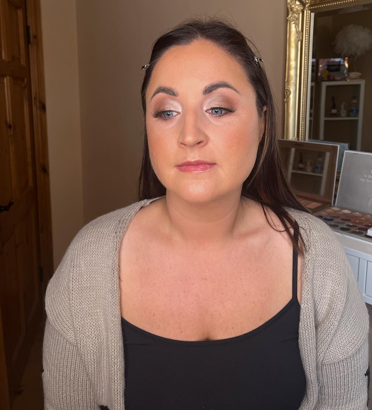 A woman with dark hair and makeup, wearing a black top and beige cardigan, in a room with a gold-framed mirror and shelves in the background. Wedding makeup in Dingle, Kerry