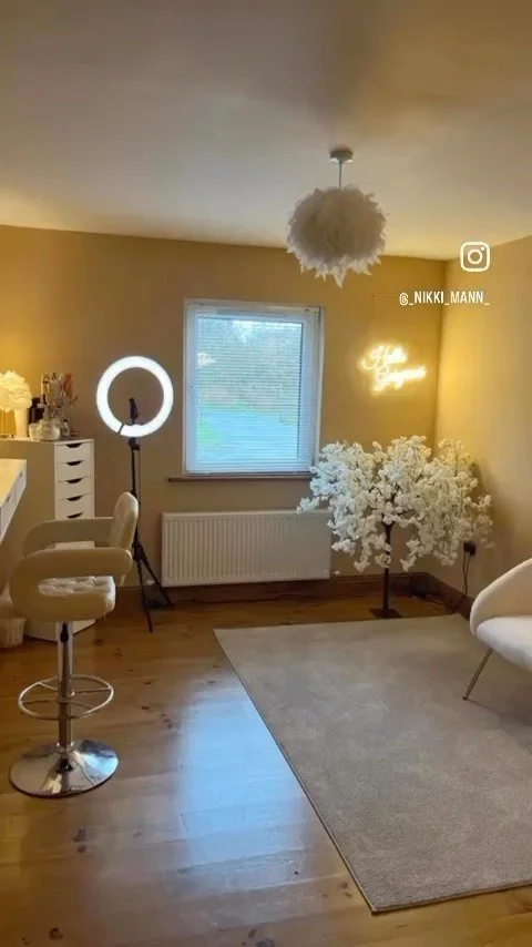 A cozy interior room with a makeup chair and ring light, a window, a white floral decorative tree, and a wall neon sign that says 'Hello Beautiful'.