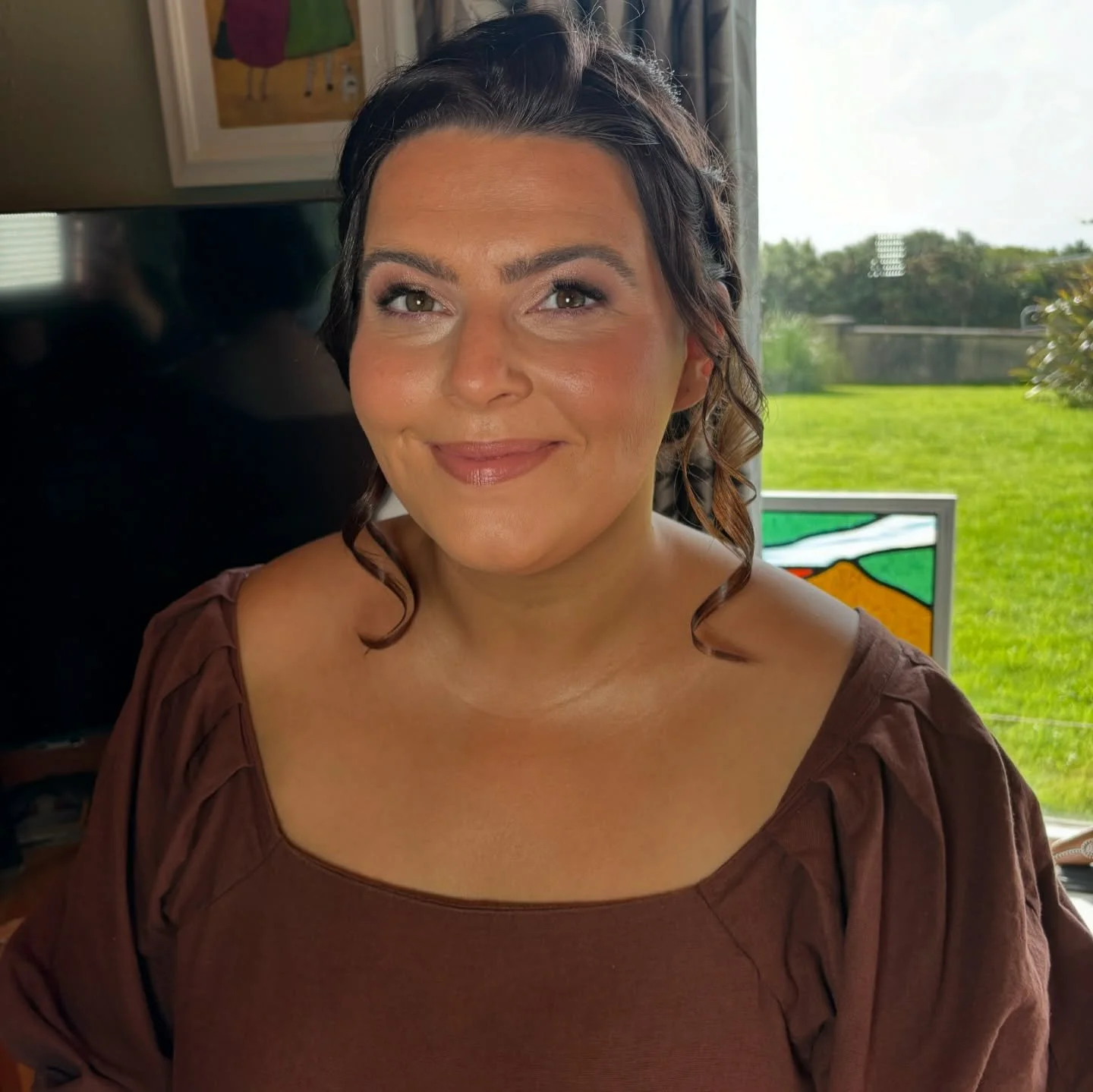 Close-up of a smiling woman with brown hair and wearing a brown top, sitting indoors near a window with a view of green grass and trees outside.
