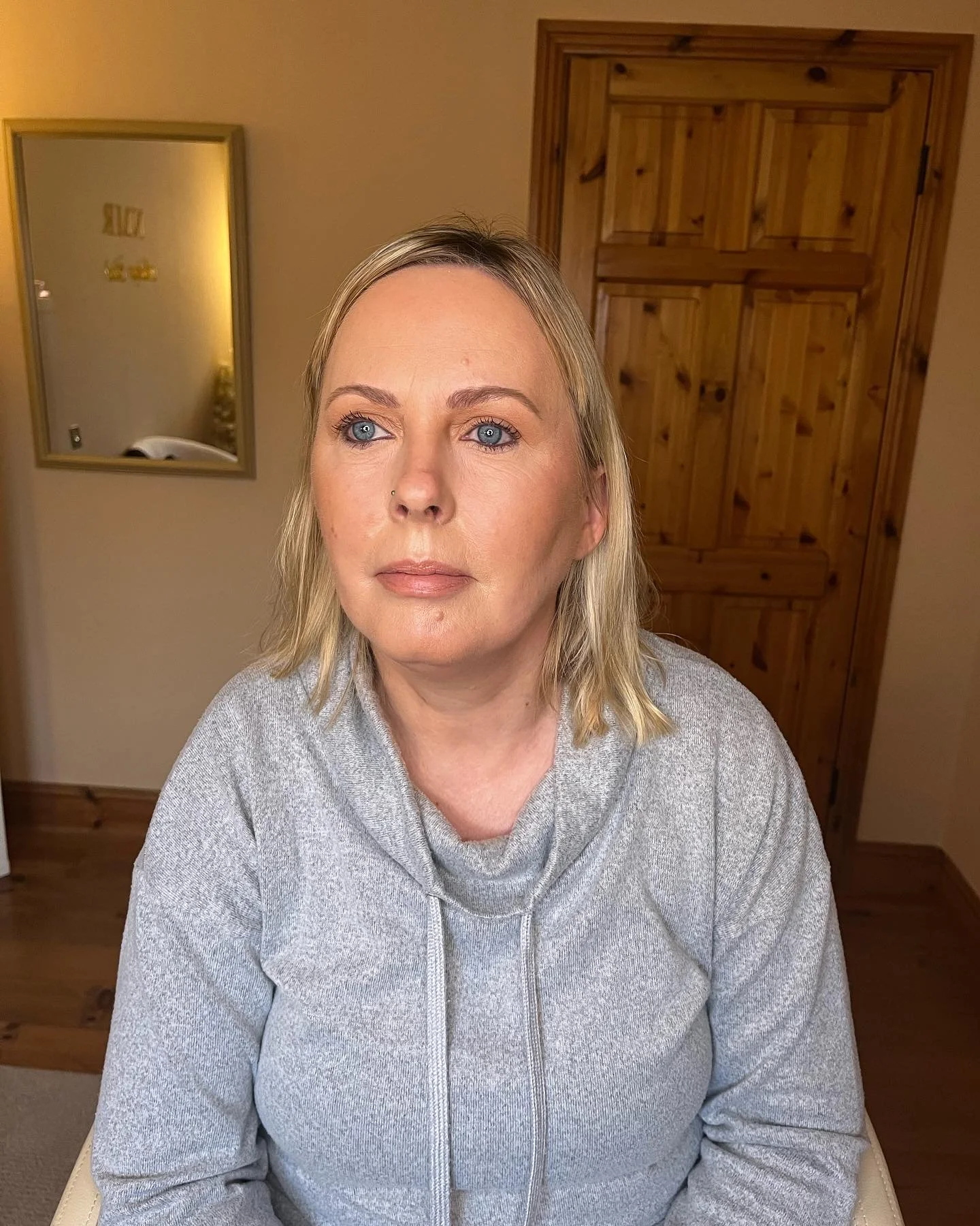 A woman with blonde hair and blue eyes sitting indoors, wearing a gray hoodie, looking slightly to the left of the camera.