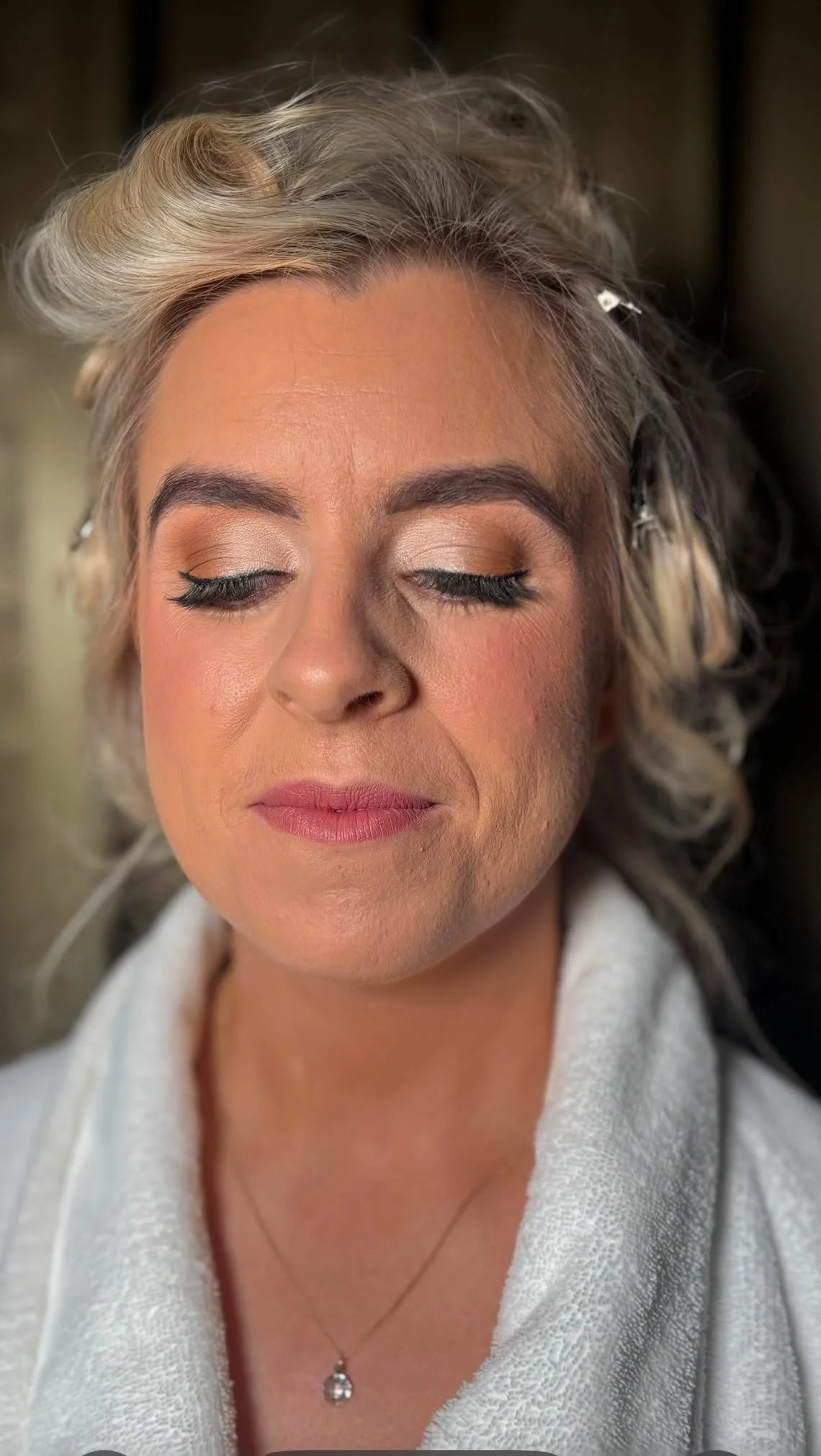 A woman with blonde hair styled in soft curls, wearing makeup with neutral eyeshadow, eyeliner, mascara, blush, and pink lipstick. She has a necklace with a pendant and is dressed in a light-colored garment with a zipper.