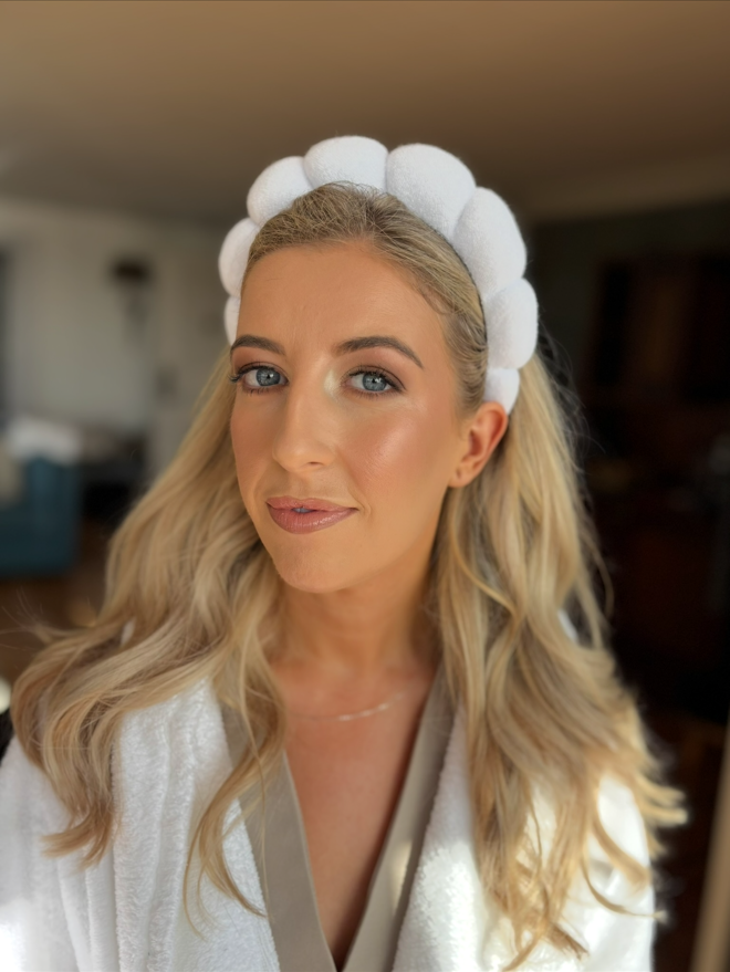 A woman with long blonde hair wearing a white headband with pink and white balls, and a white bathrobe, smiling in a home setting.
