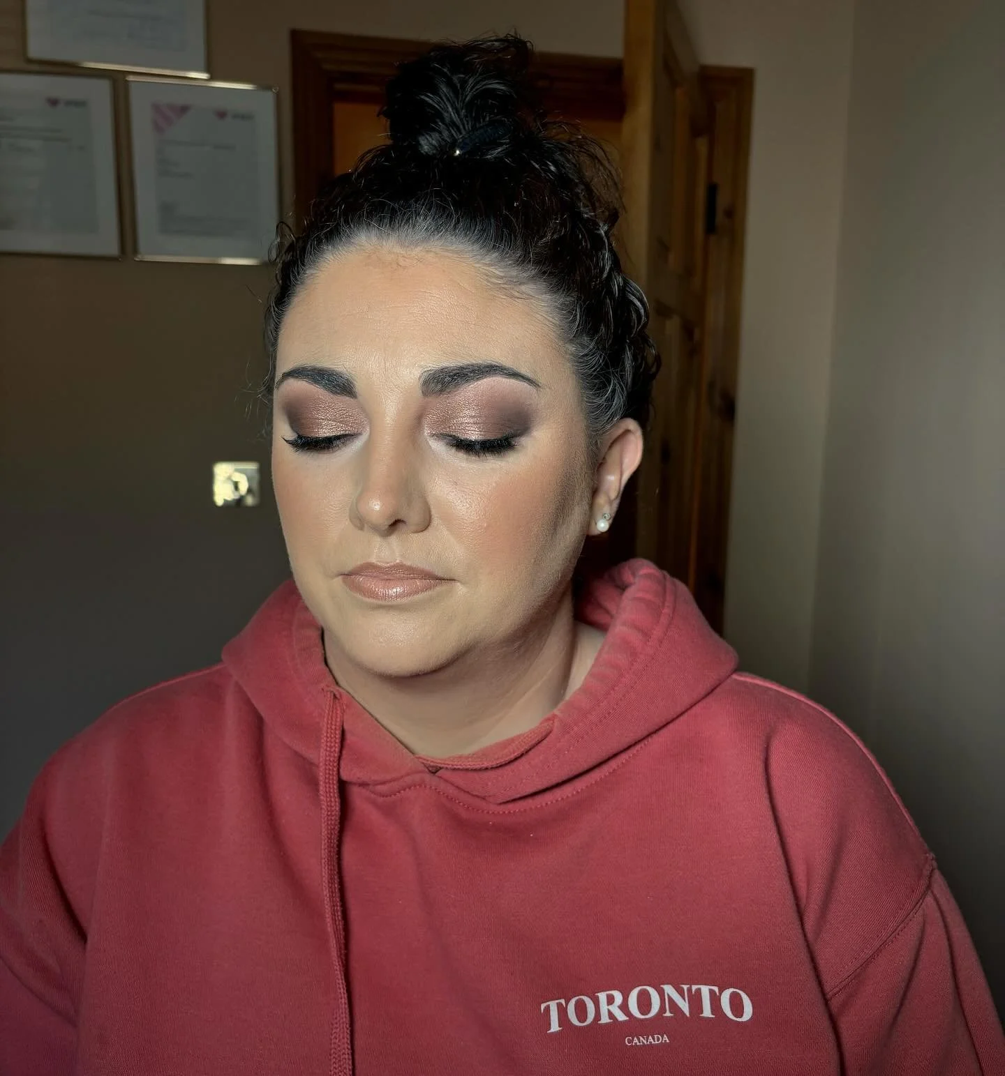 A woman with closed eyes, wearing makeup and pearl earrings, in a red hoodie with 'Toronto Canada' written on it, standing indoors with a wooden door and framed pictures in the background.