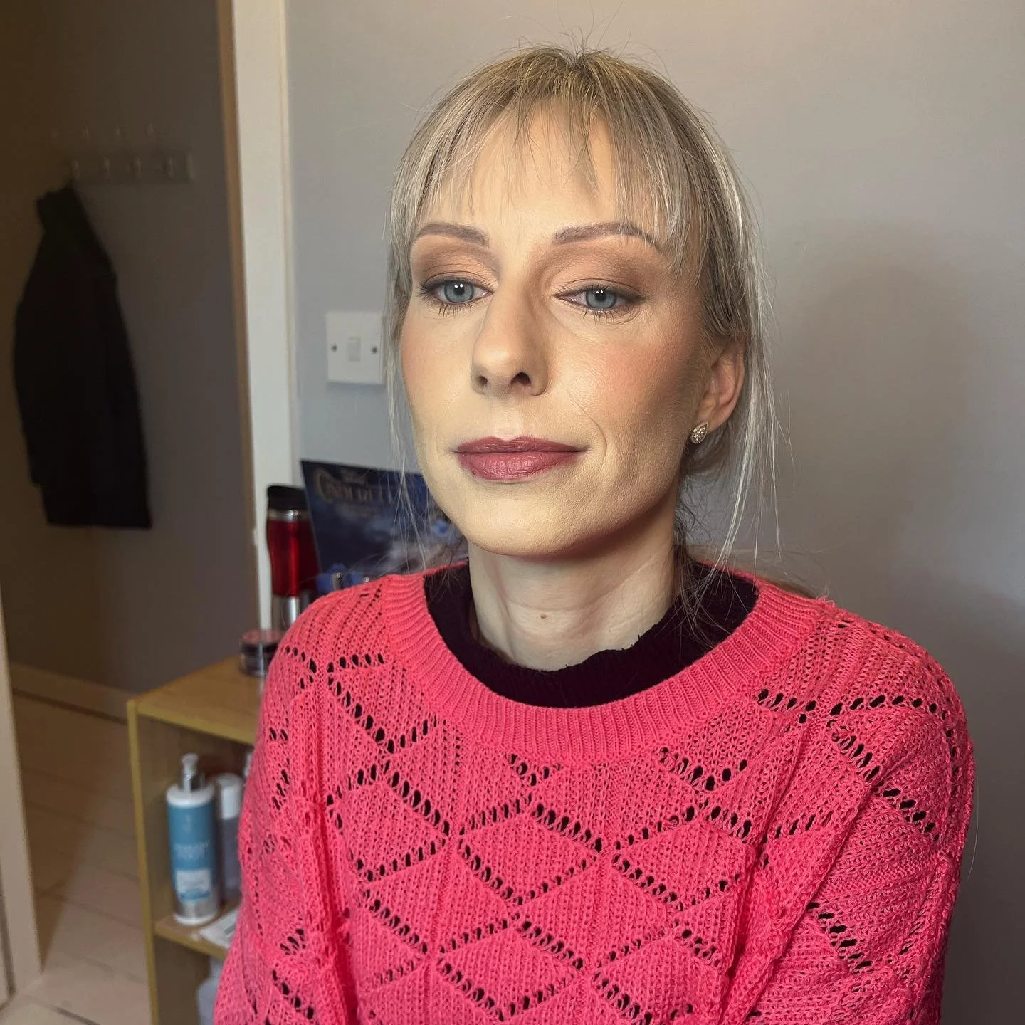 A woman with blonde hair and blue eyes wearing a pink knitted sweater, standing indoors with a neutral wall background.