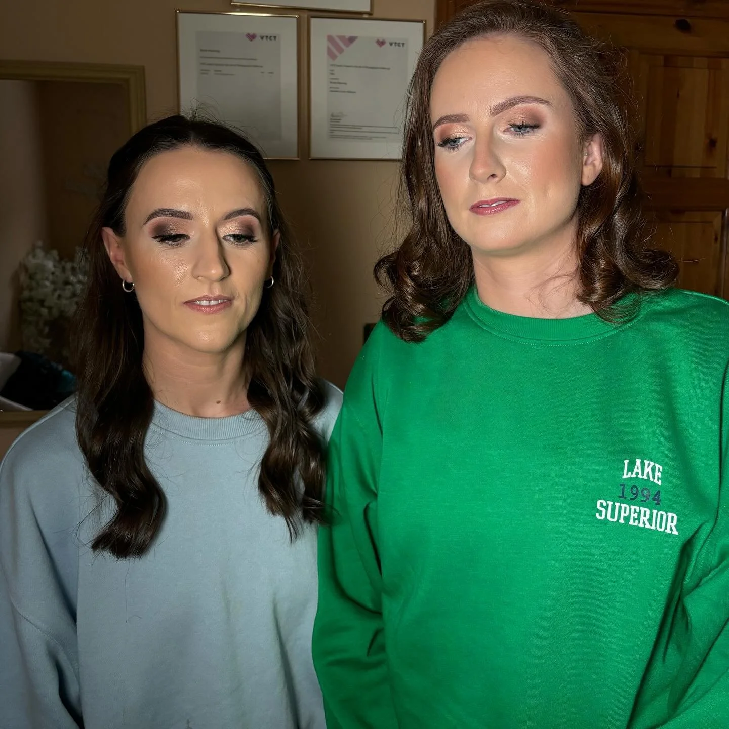 Two women standing together, one wearing a light gray sweatshirt and the other wearing a green sweatshirt with text, both with closed eyes and neutral expressions, in a room with framed certificates on the wall.