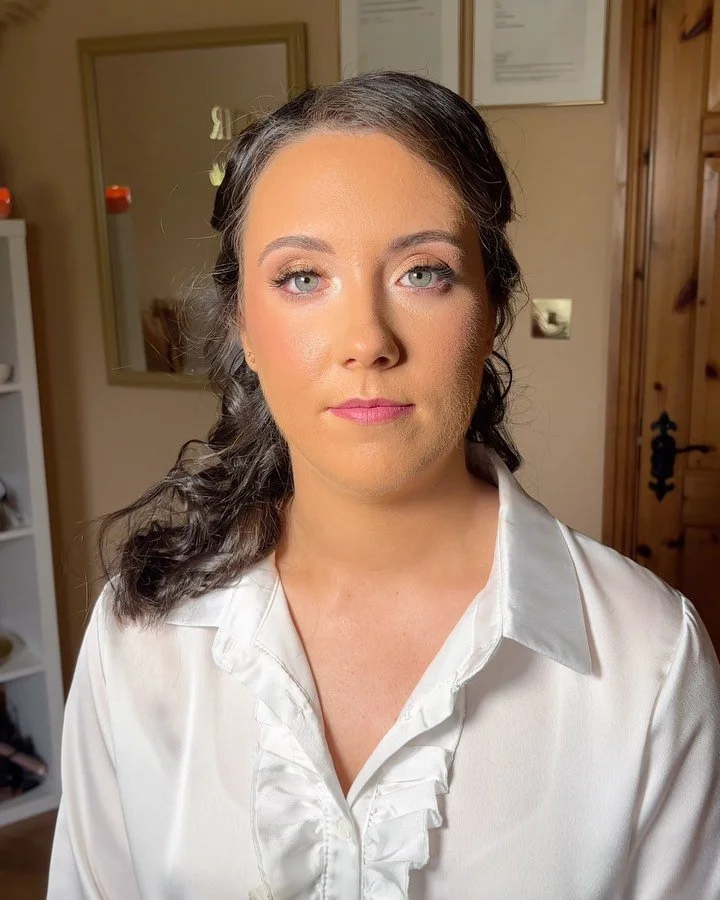 An Irish woman with long dark hair styled in loose curls, wearing a white satin blouse, looking directly at the camera in a room with framed pictures and a wooden door in the background.
