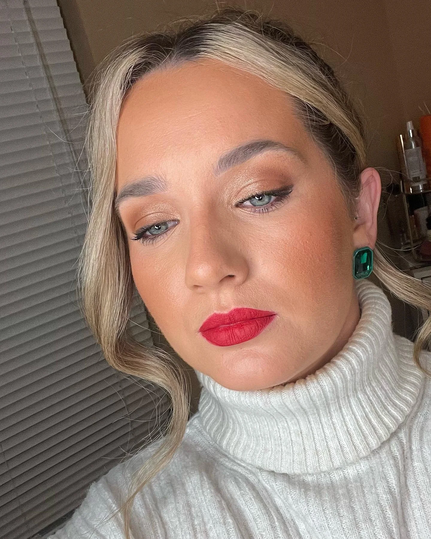 Close-up of a woman with blonde, wavy hair, blue eyes, wearing red lipstick, gold eyeshadow, and teal earrings, dressed in a white turtleneck sweater.