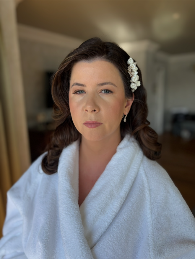 A woman with dark brown hair styled in loose waves, wearing a white robe and a floral hair accessory, standing indoors with a neutral expression.