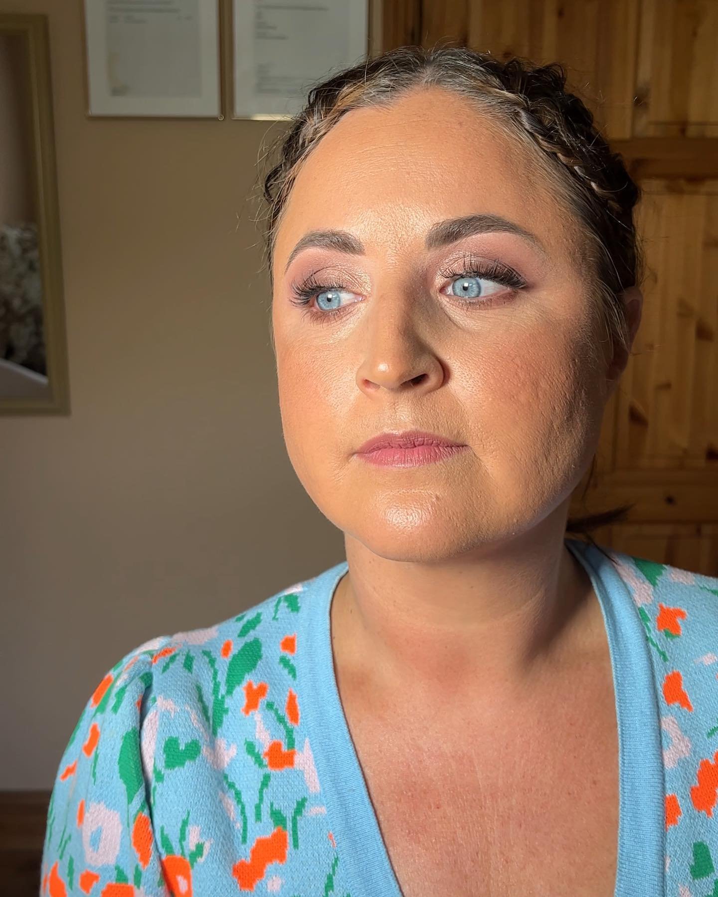 Close-up of a woman with blue eyes and makeup, wearing a blue shirt with orange and green floral print, standing in a room with wooden walls.
