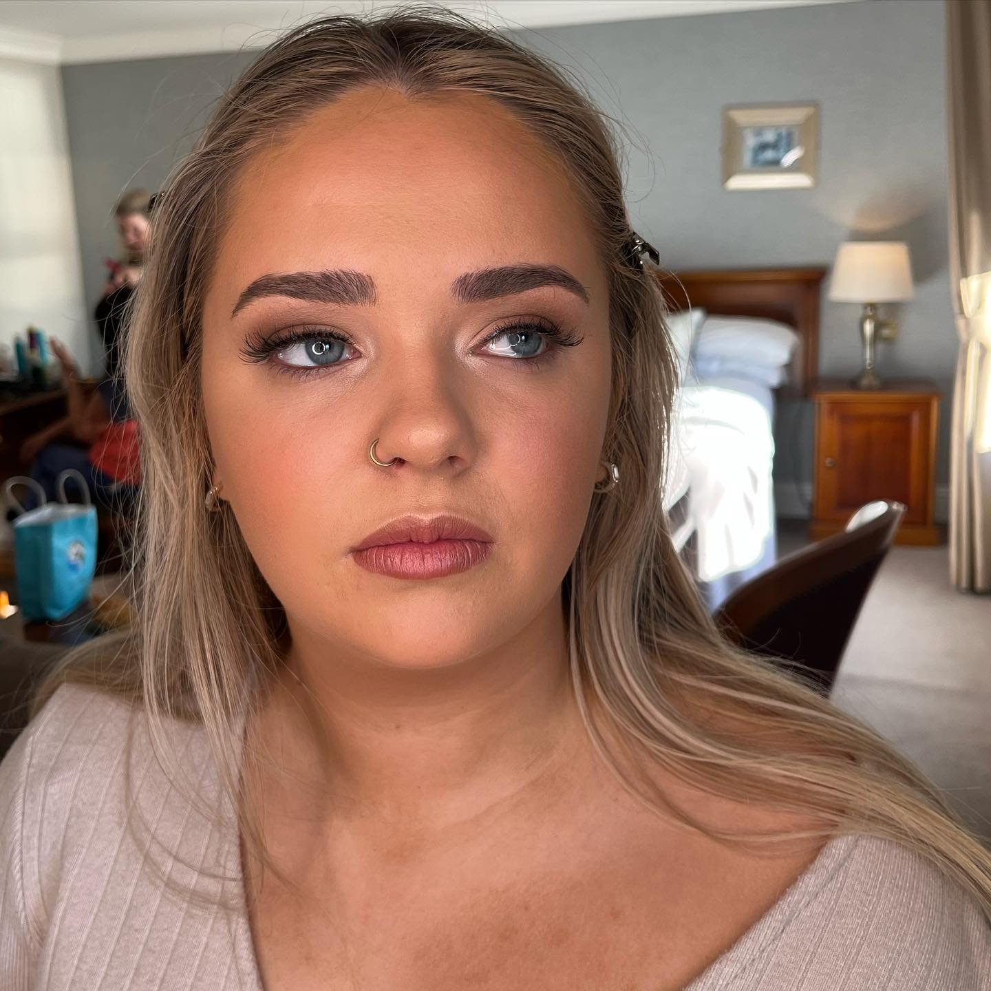 Close-up of a woman with makeup, light skin, blonde hair, wearing a beige top, and a small nose ring, in a hotel room with a bed, bedside lamps, and a mirror in the background.