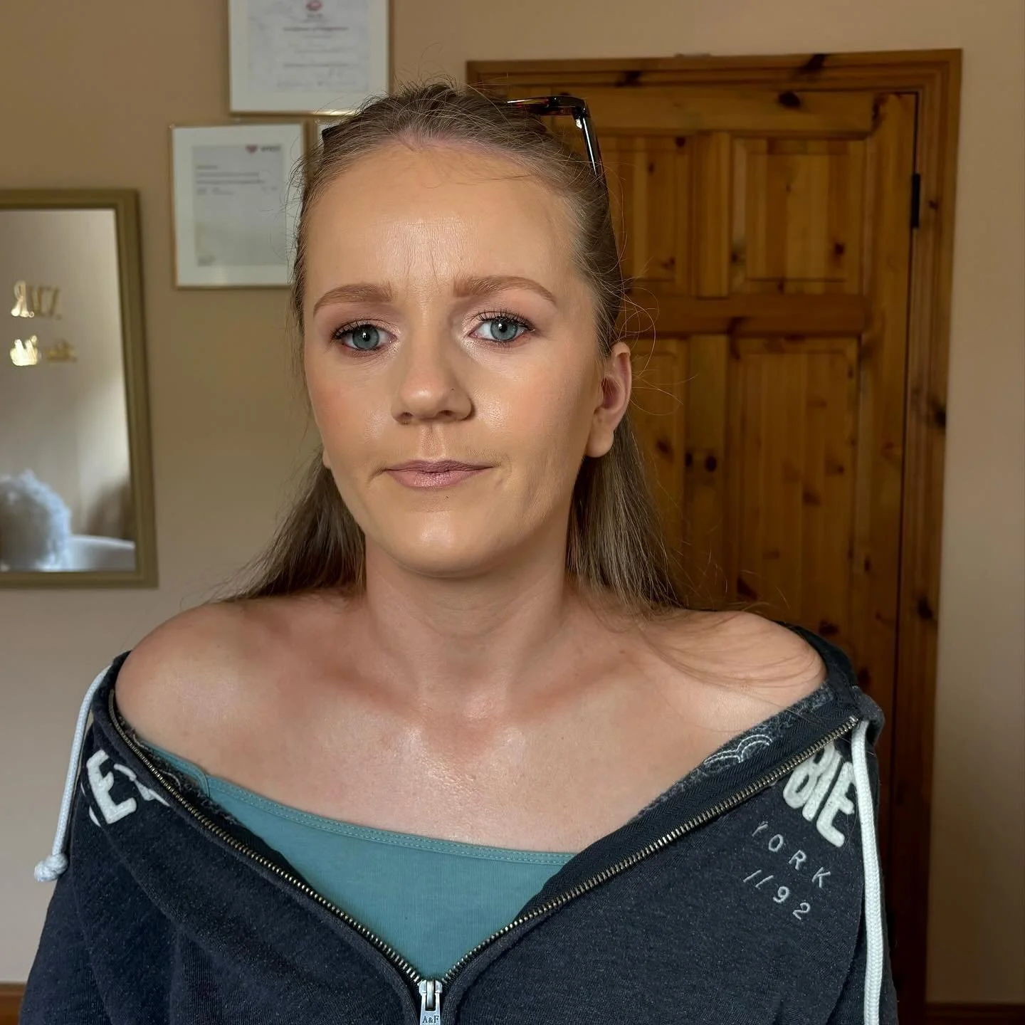 A woman with light skin, long brown hair, and blue eyes standing indoors in front of a wooden door and framed documents on the wall, wearing a dark hoodie with a zipper and an off-the-shoulder top underneath.
