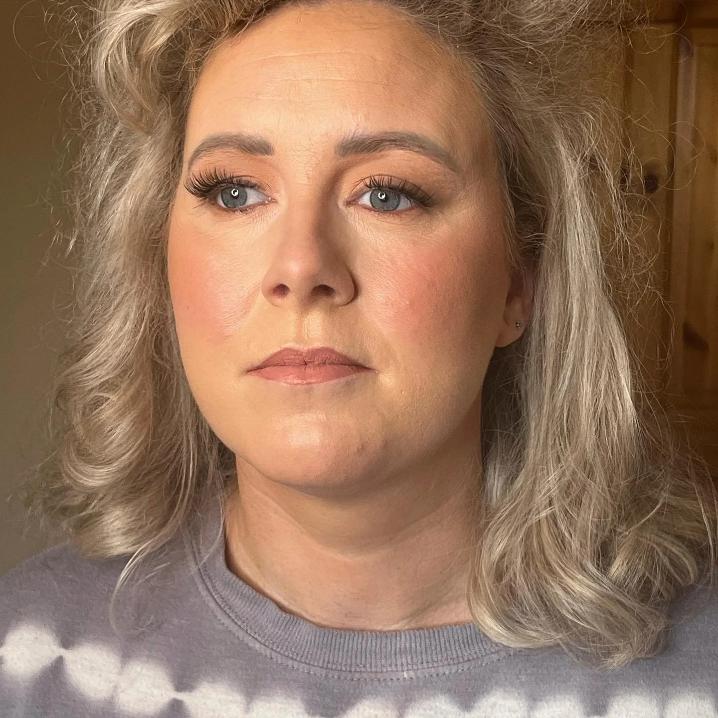 Close-up of a middle-aged woman with curly, shoulder-length blonde hair and blue eyes, looking slightly to the side, wearing a gray sweater with a pattern, in a room with wood-paneled background.