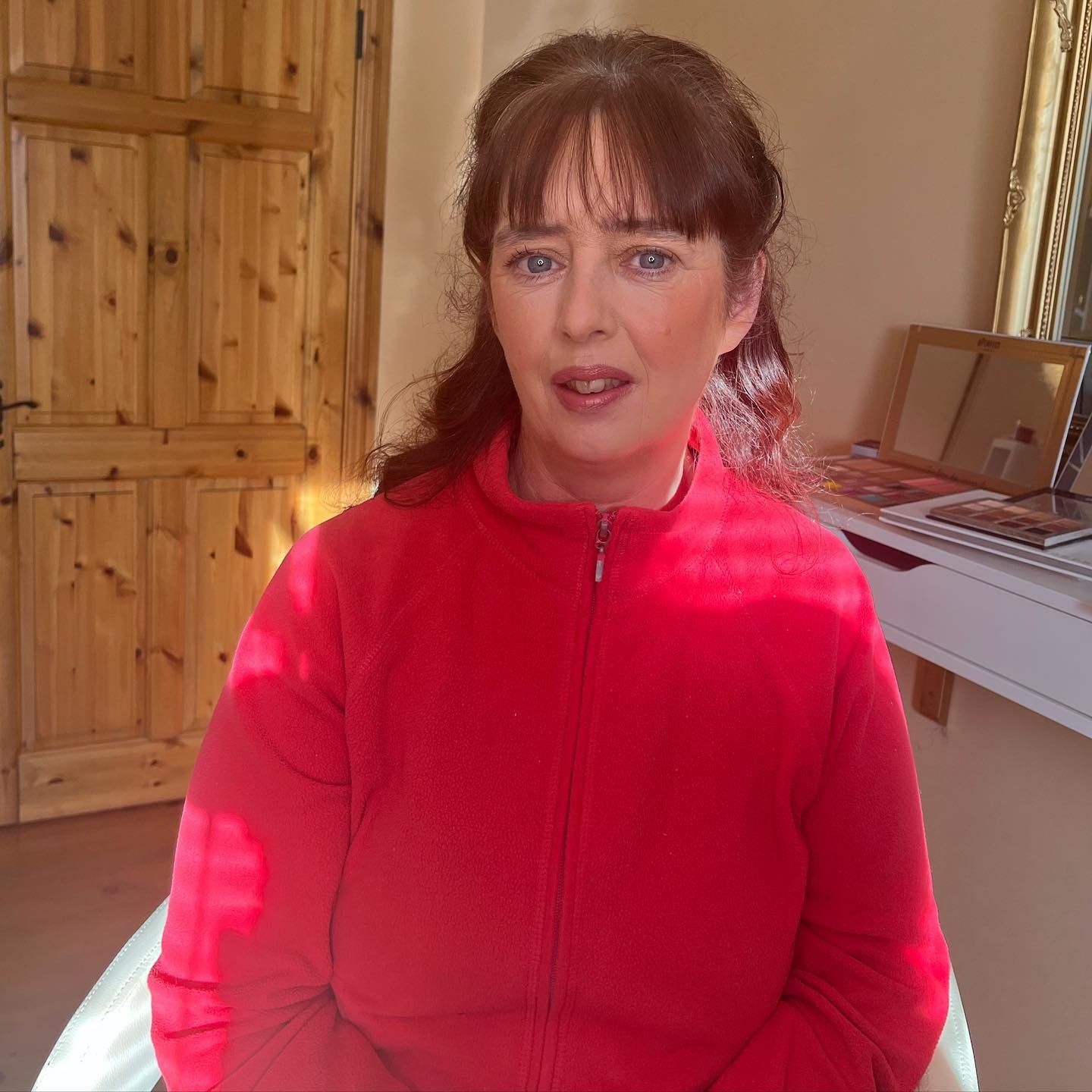 A woman with brown hair and bangs wearing a red zip-up jacket, sitting indoors in front of a wooden door and a mirror on a white table.