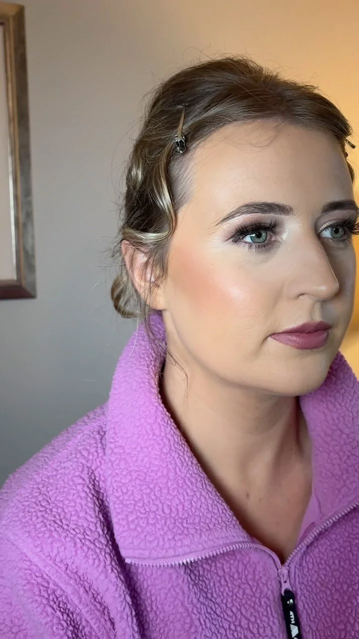 Close-up of a woman with styled hair and makeup, wearing a purple fleece jacket, in an indoor setting.