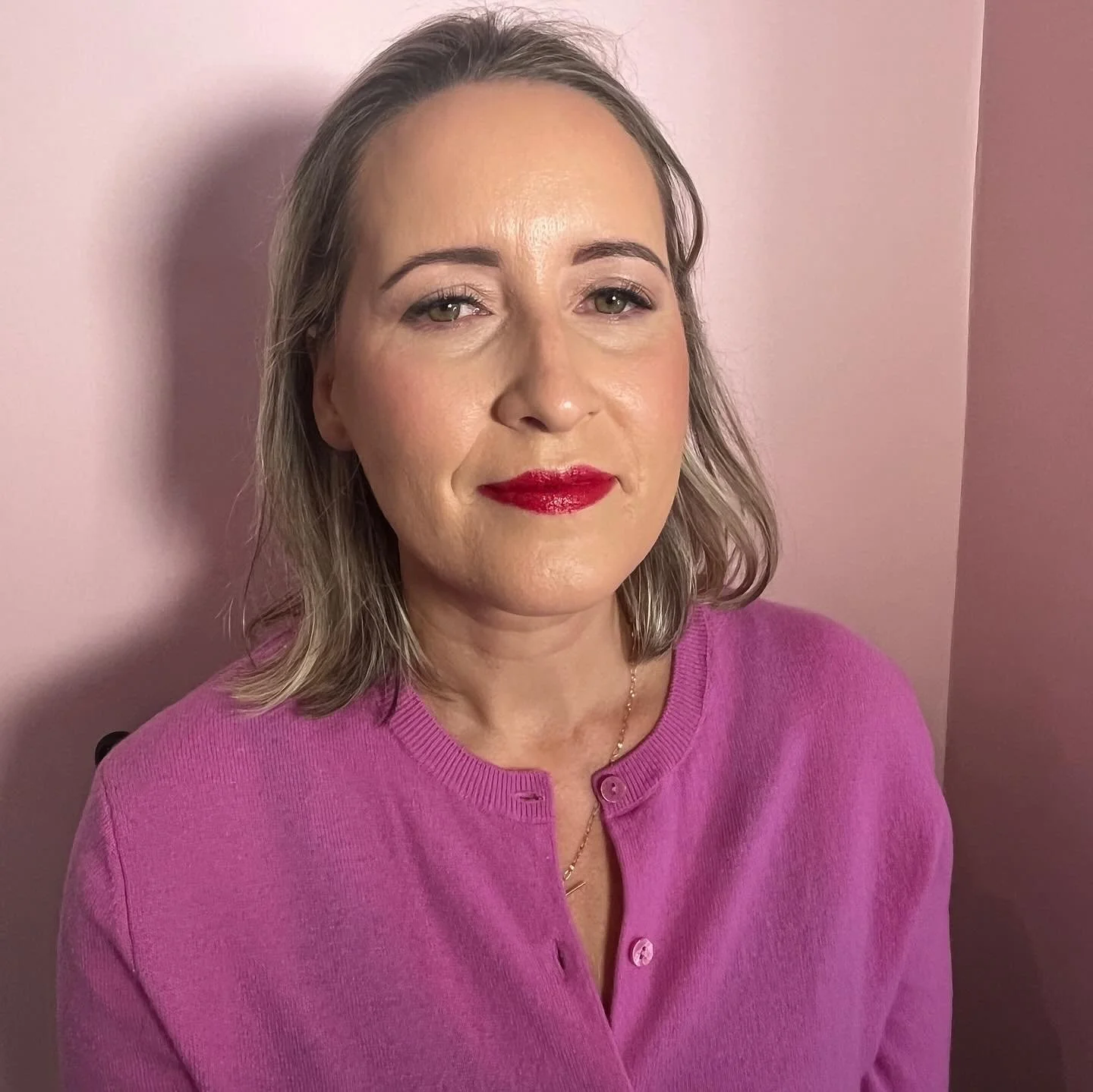 A woman with shoulder-length light brown hair wearing a pink top and red lipstick, standing against a pink wall.