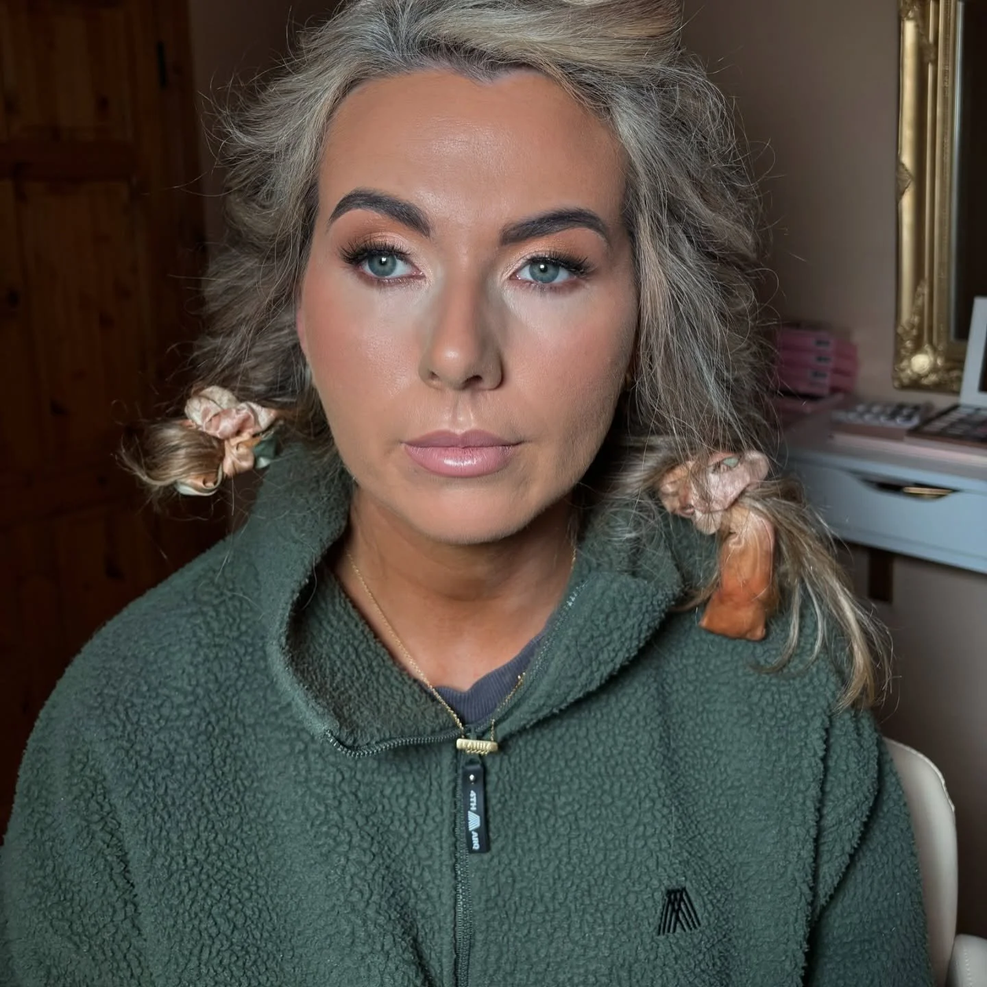 A woman with curly hair styled with pink scrunchies, wearing a green fleece jacket, sitting indoors.