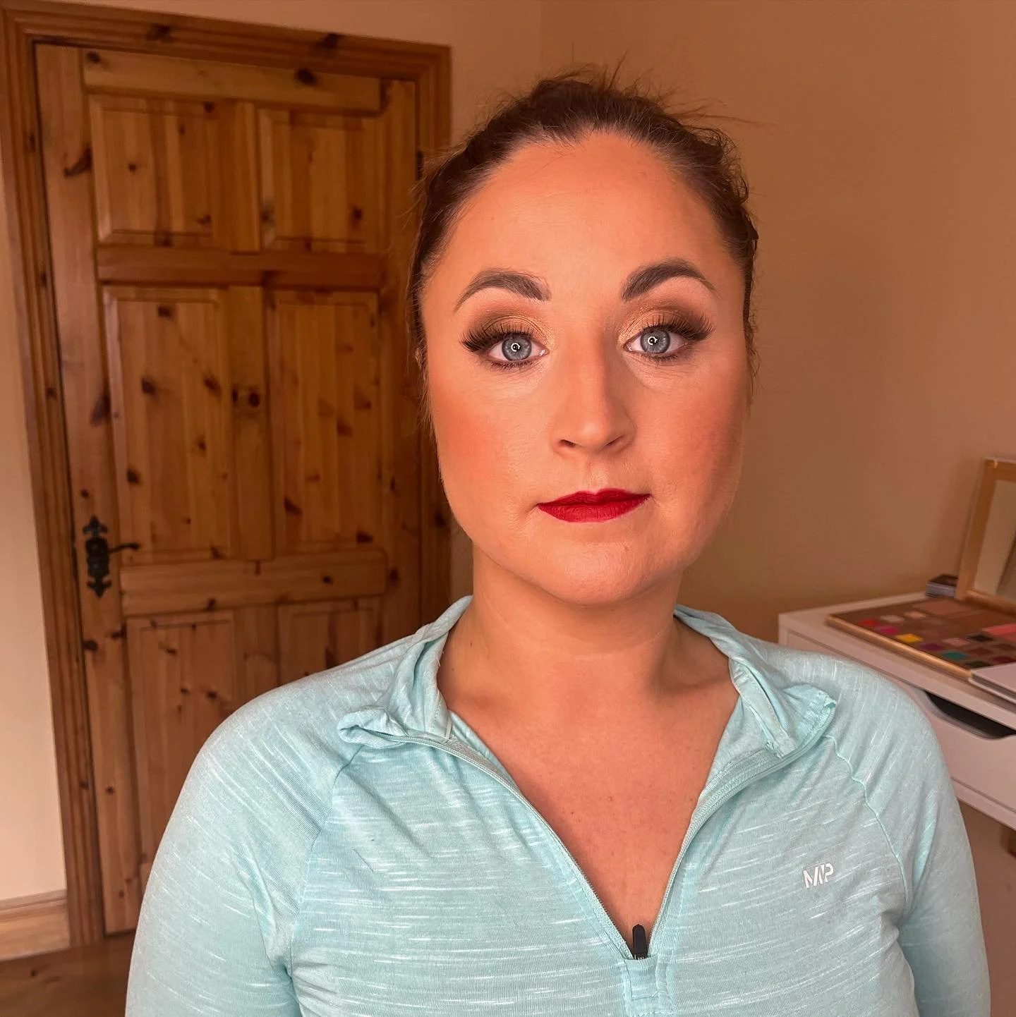 A woman with makeup, including dark eye makeup and red lipstick, wearing a light blue athletic top in a room with wooden doors and a makeup palette on a desk.