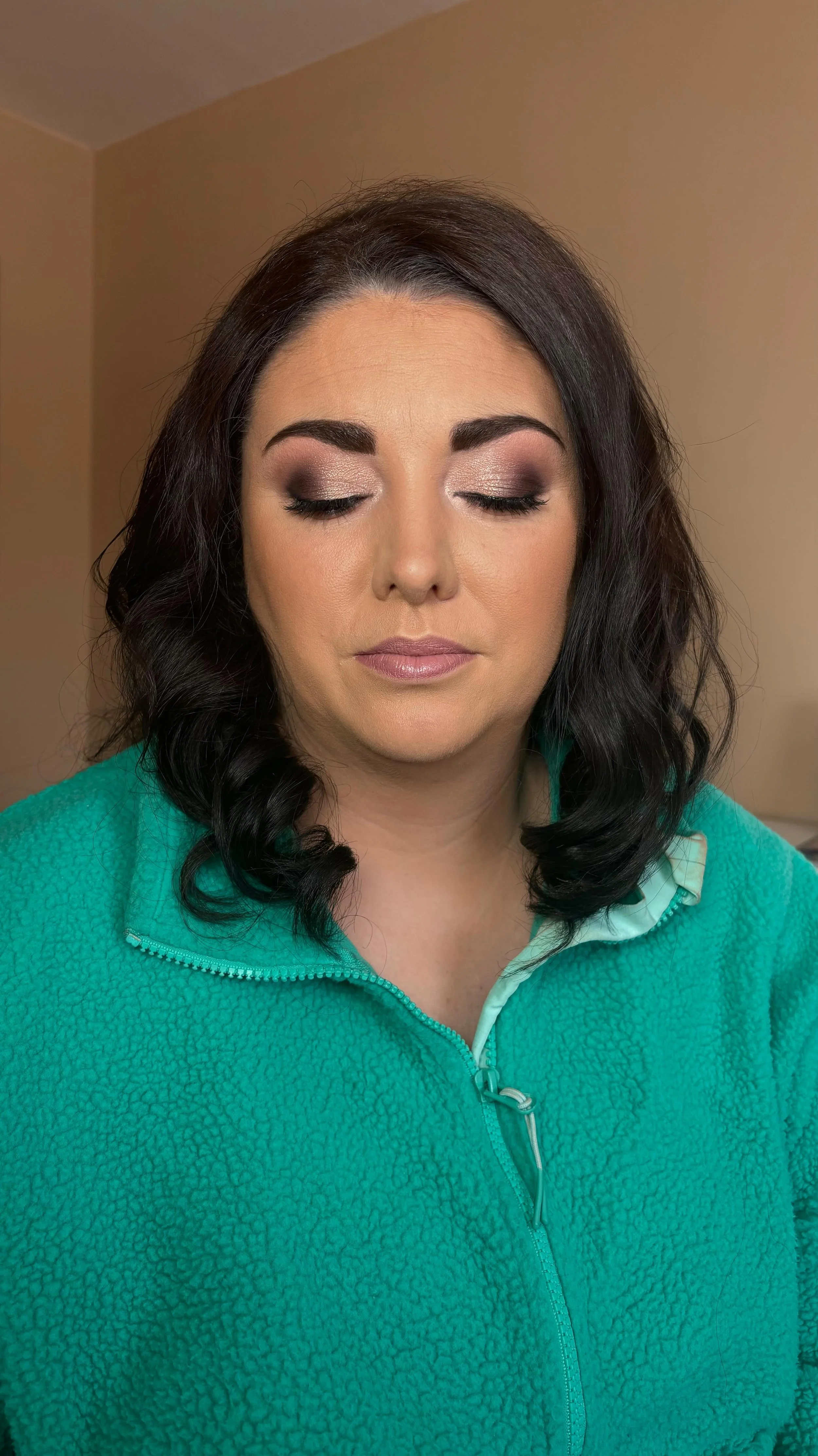 A woman with dark, wavy hair and makeup, wearing a teal fleece jacket with a zipper, closing her eyes.