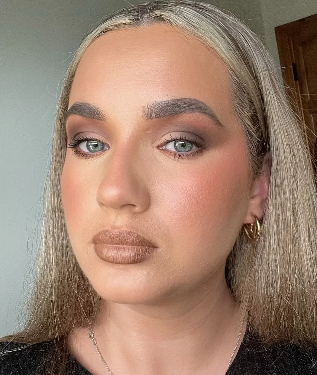 Close-up of a woman with blonde hair, blue eyes, wearing gold hoop earrings, subtle makeup, and a black top.