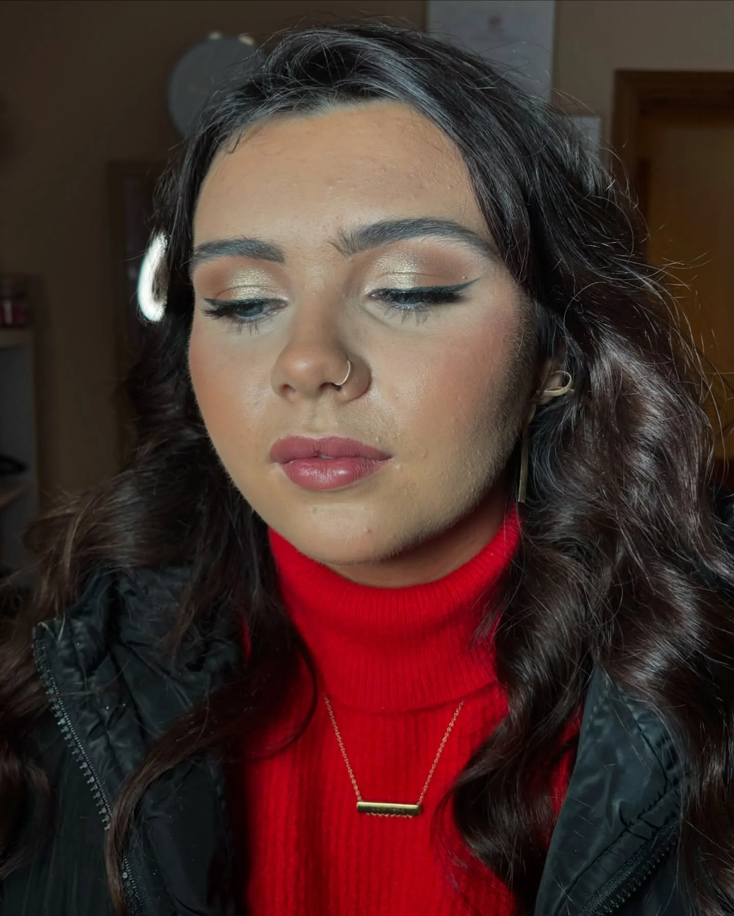 A woman with dark wavy hair, wearing a red turtleneck sweater, black jacket, and gold jewelry, looking downward with makeup including gold eyeshadow and winged eyeliner, indoor background.