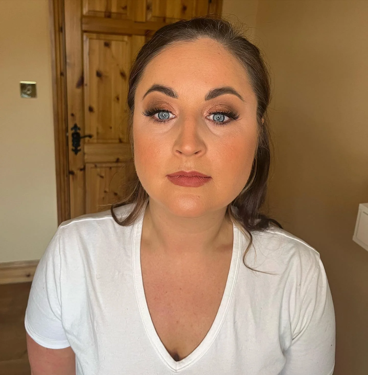 A woman with light skin, brown hair, and blue eyes, wearing makeup, a white shirt, and looking slightly to the left, standing in a room with wooden furniture and beige walls.