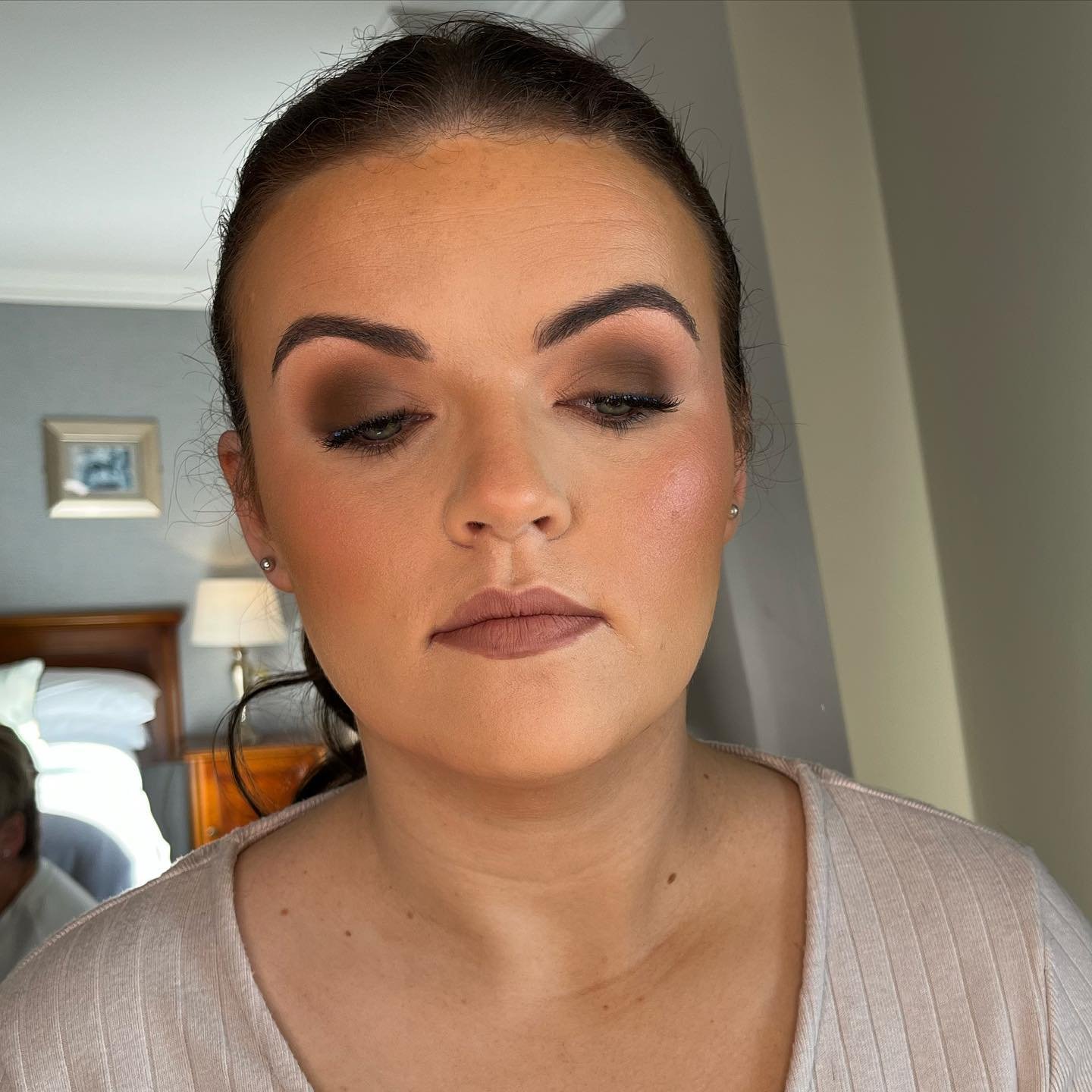 A woman with dark hair pulled back, wearing makeup, a beige top, and small earrings, standing in a room with a bed, lamp, and wall decorations in the background.