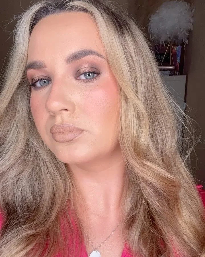 Close-up of a woman with long, wavy blonde hair, wearing makeup, and a pink top, posing indoors with a blurred background.