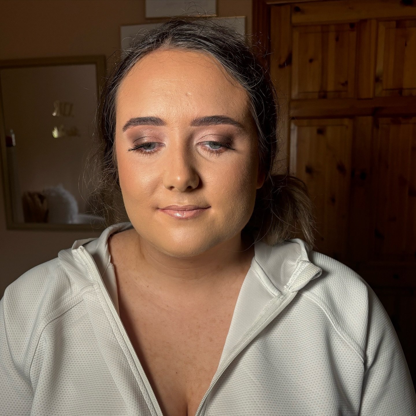 A woman with medium-length dark hair tied back, wearing makeup, and a light-colored zip-up jacket, sitting indoors with a wooden cabinet and mirror in the background.