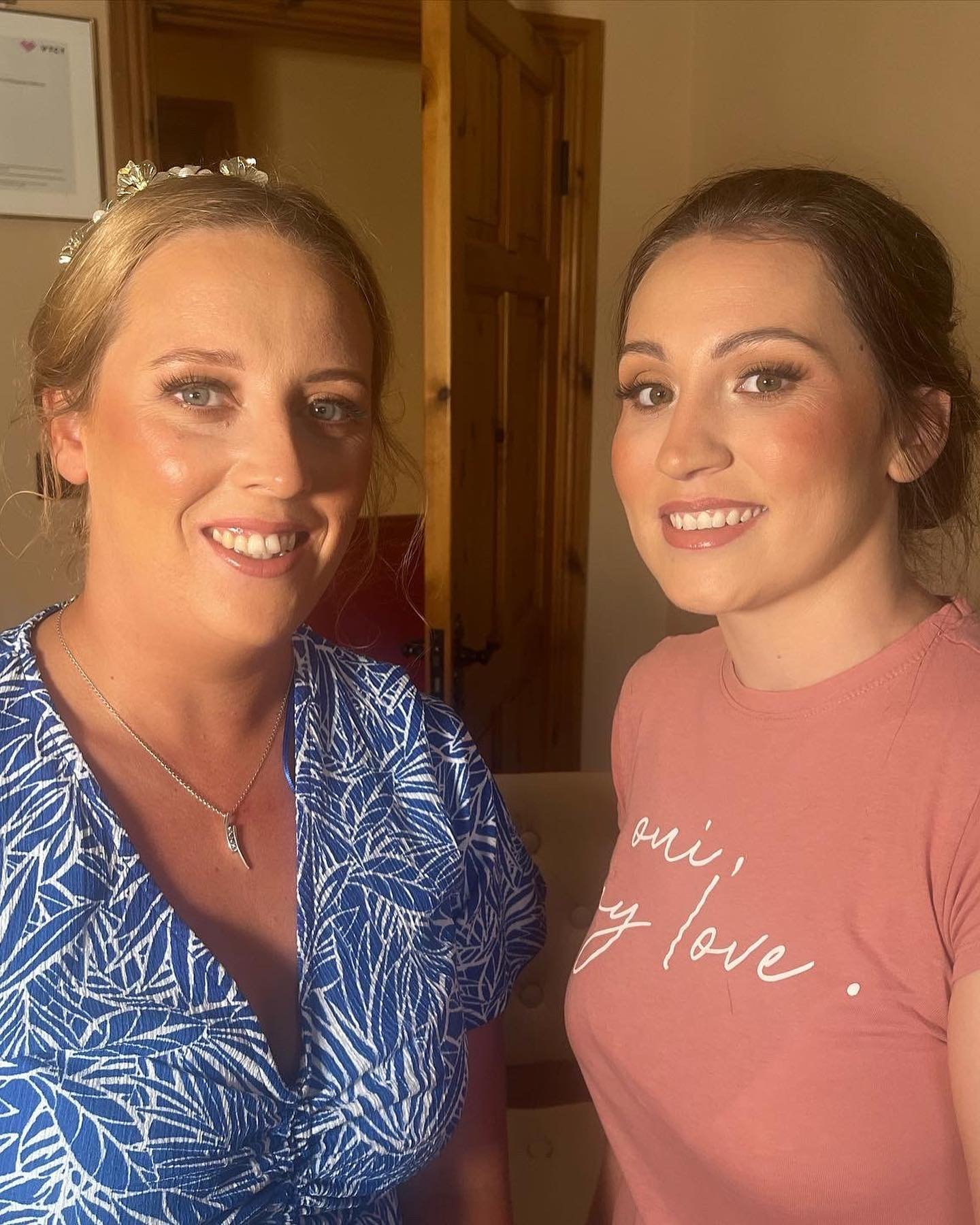 Two women smiling for a selfie indoors. The woman on the left has blonde hair, blue eyes, wears a blue and white patterned shirt, a necklace, and flowers in her hair. The woman on the right has light brown hair, sea-green eyes, and wears a pink shirt