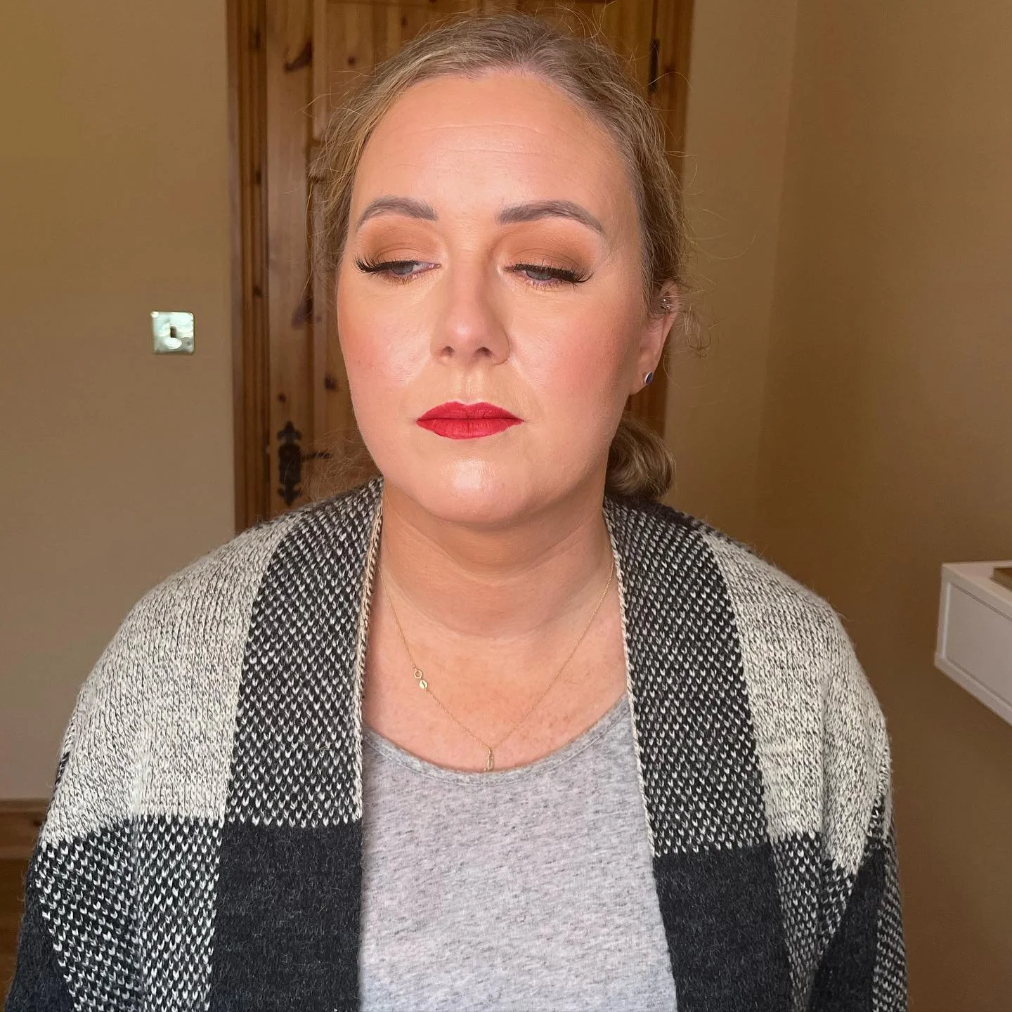 A woman with blonde hair styled in a loose bun, wearing makeup with winged eyeliner and red lipstick, dressed in a light gray top and black-and-white checkered cardigan, standing indoors with a wooden door and beige wall in the background in Dingle