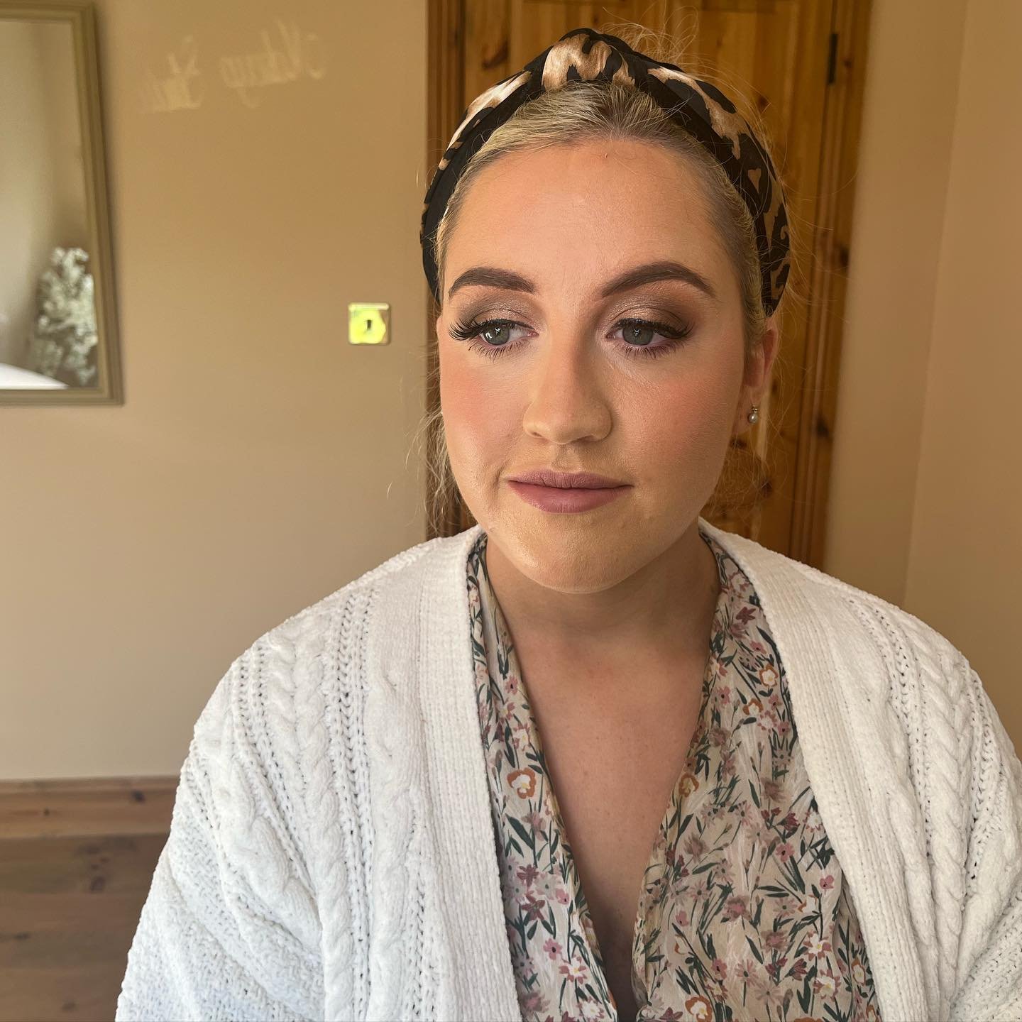 Close-up of a woman with blonde hair wearing a patterned headband, makeup, a floral blouse, and a white knitted cardigan, standing indoors in a room with a mirror and wooden furniture.