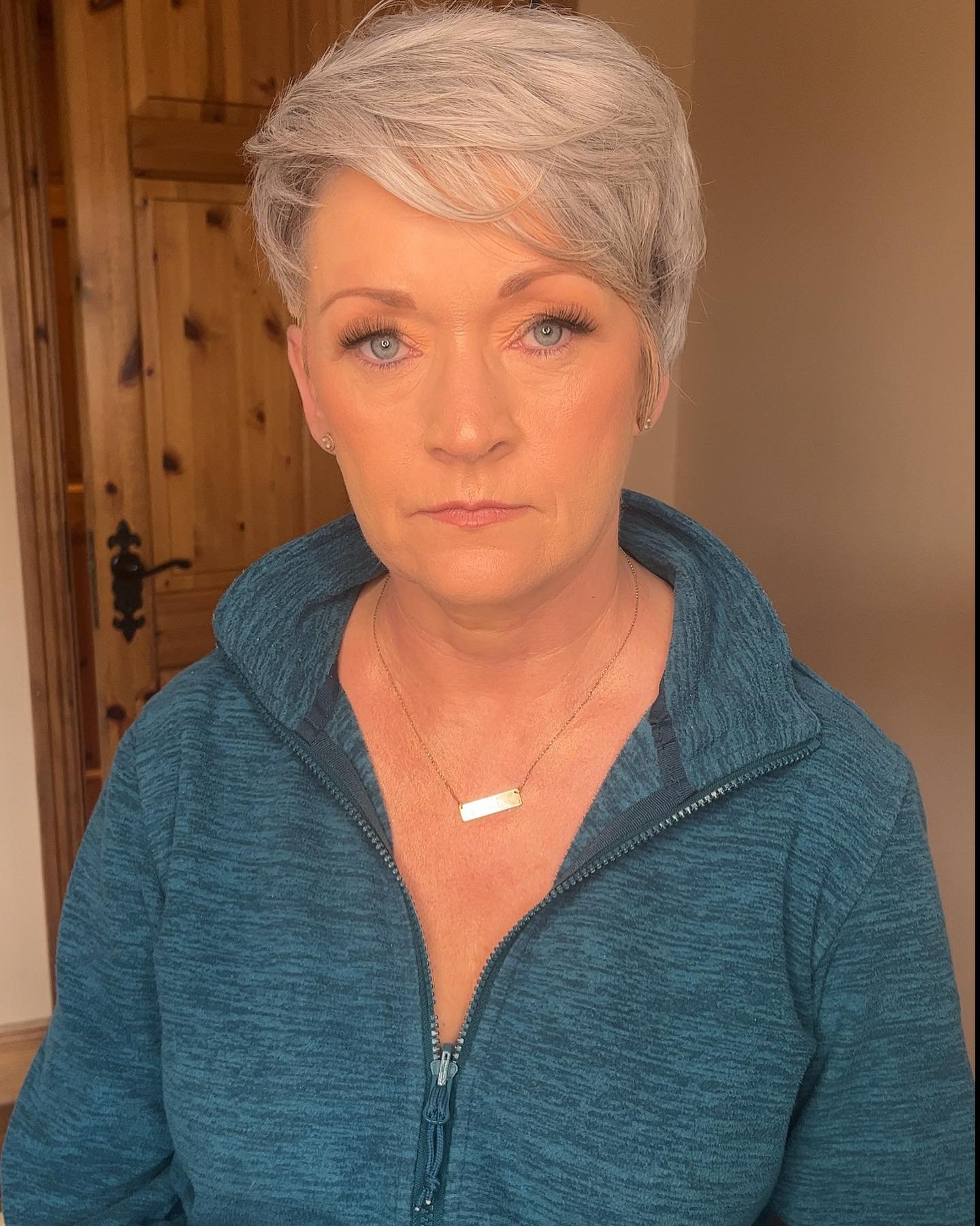A woman with short gray hair, wearing a blue zip-up jacket, a gold necklace, and earrings, standing indoors in front of a wooden door.