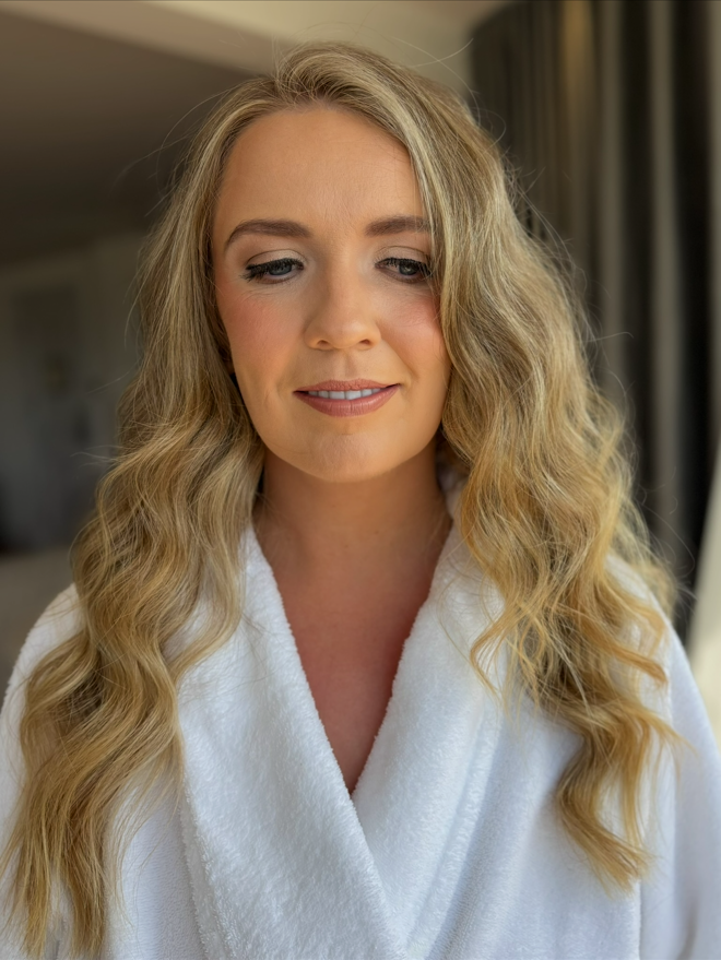 Close-up of a woman with wavy blonde hair, wearing a white robe, looking down with a gentle smile.