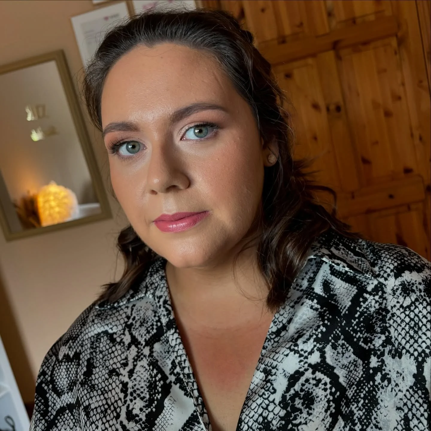 A woman with brown hair and blue eyes wearing makeup, a patterned black and white top, in a room with wooden walls and a mirror reflecting a lamp and a framed picture.