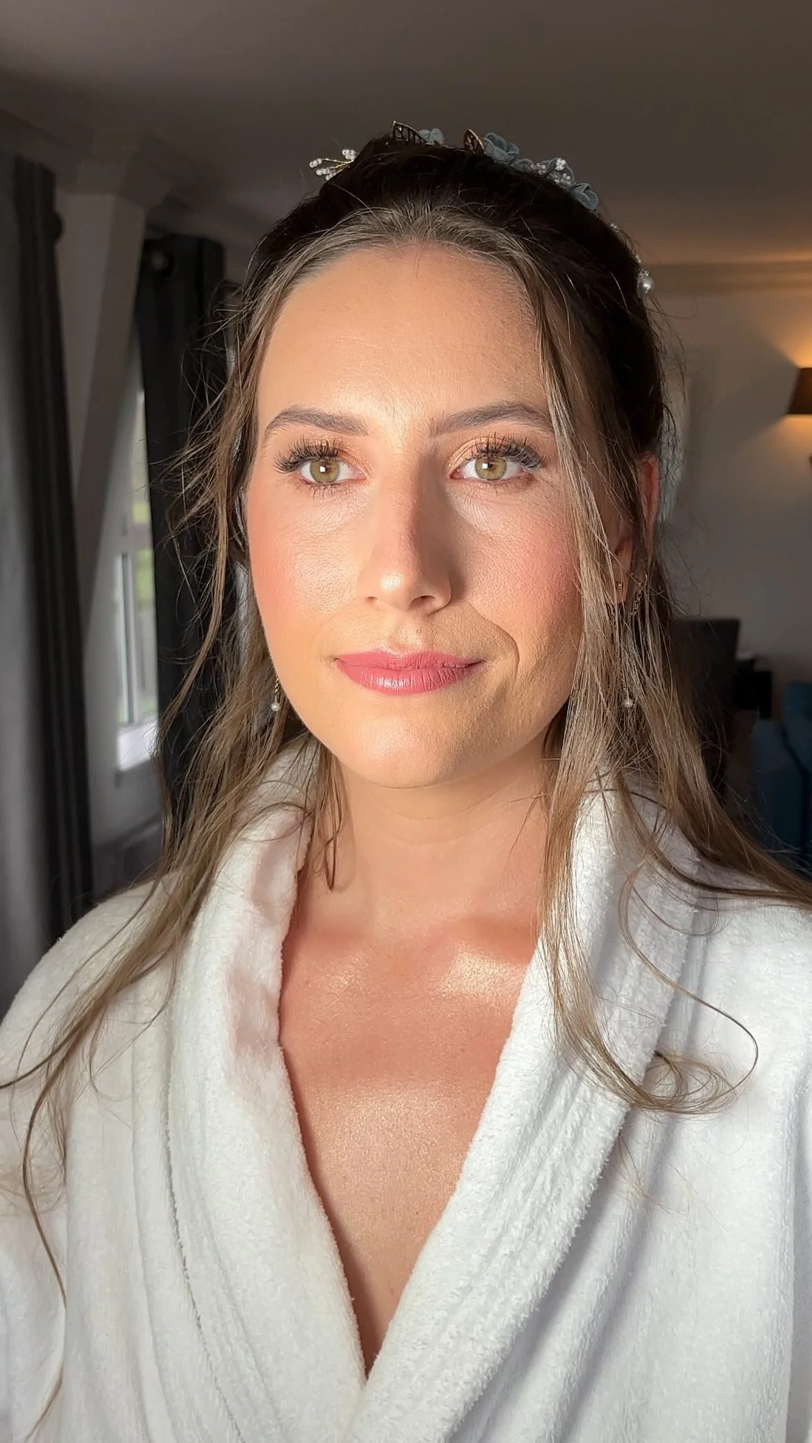 A woman with long, wavy brown hair and hazel eyes, wearing a white bathrobe, standing near windows with dark curtains, in a well-lit room.