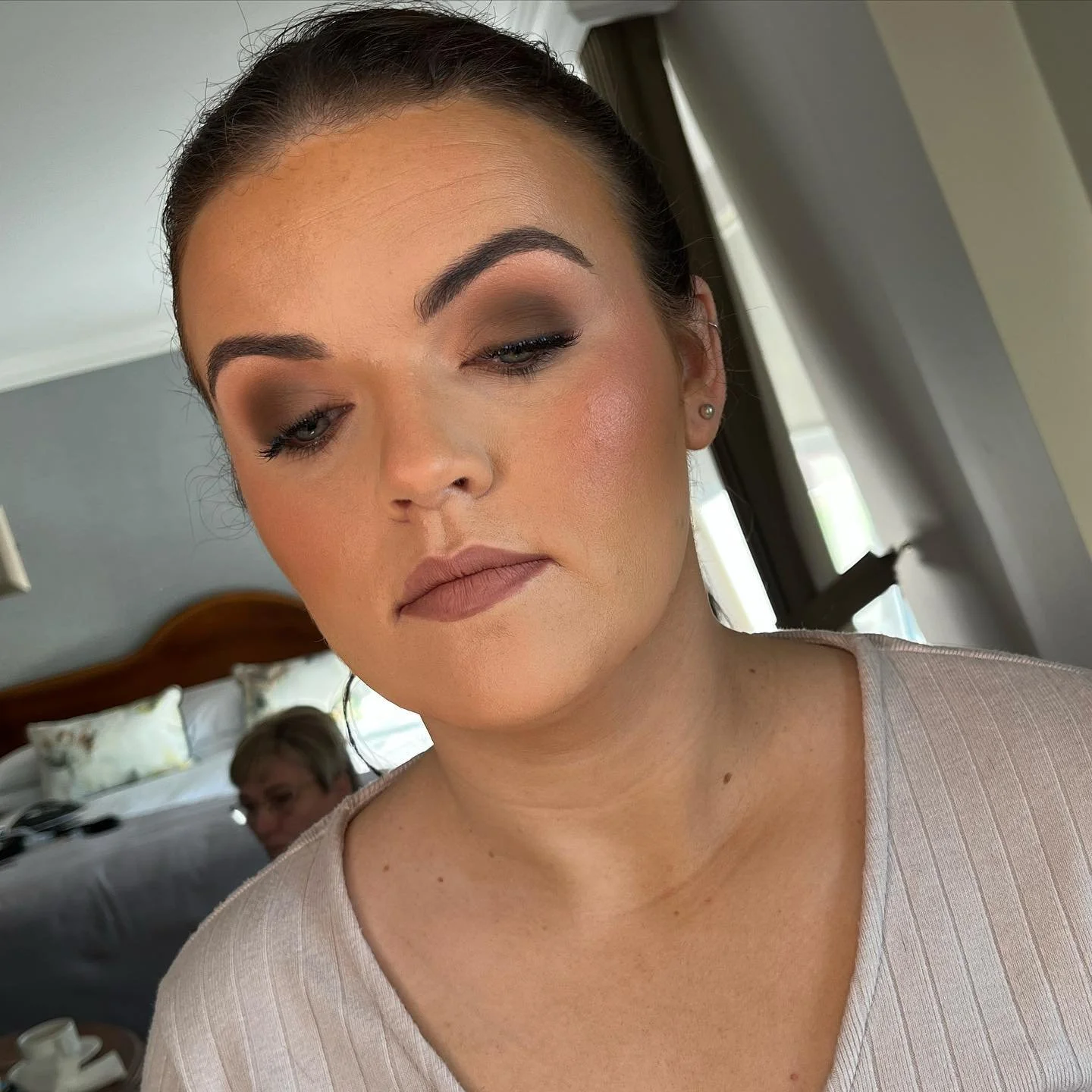 Close-up of a woman with makeup and earrings, standing near a window in a room with a bed and a person in the background.