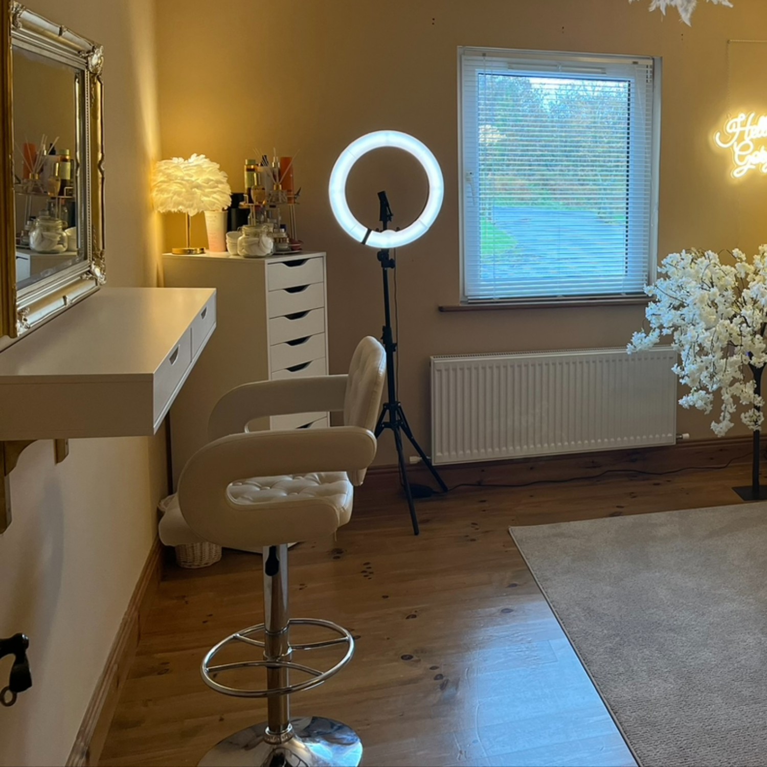 A cozy room with a makeup station, including a white chair, a ring light, a white dresser, a mirror, and decorative items like a lampshade, flowers, and a neon sign.