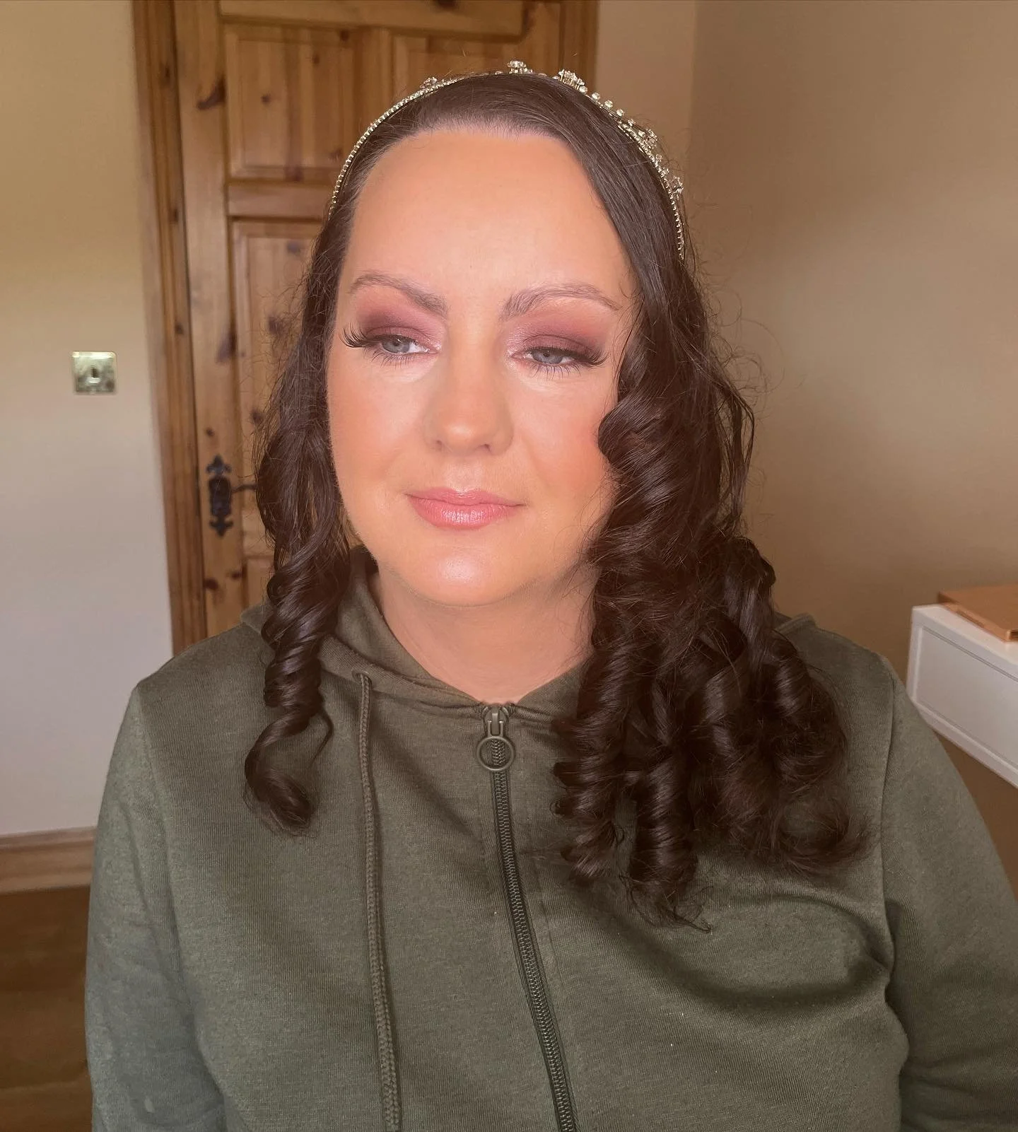 A woman with long, dark, curly hair wearing a headband and a green zip-up jacket, looking slightly to the side in a room with a wooden door and beige walls.