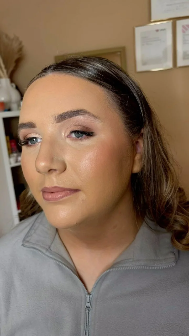 Close-up of a woman with fair skin, blue eyes, and dark brown hair styled in loose waves. She is wearing makeup with neutral tones, a soft lip color, and light eyeshadow. She is dressed in a gray zip-up top and is looking slightly to the side.